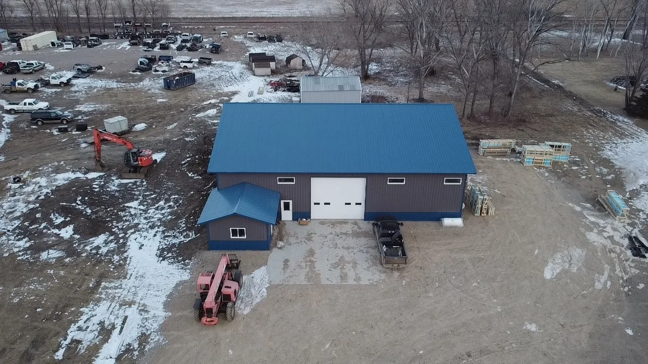 Aerial view of a large blue building with a matching smaller structure attached. Site has construction equipment, vehicles, and materials scattered around, with trees in the background.