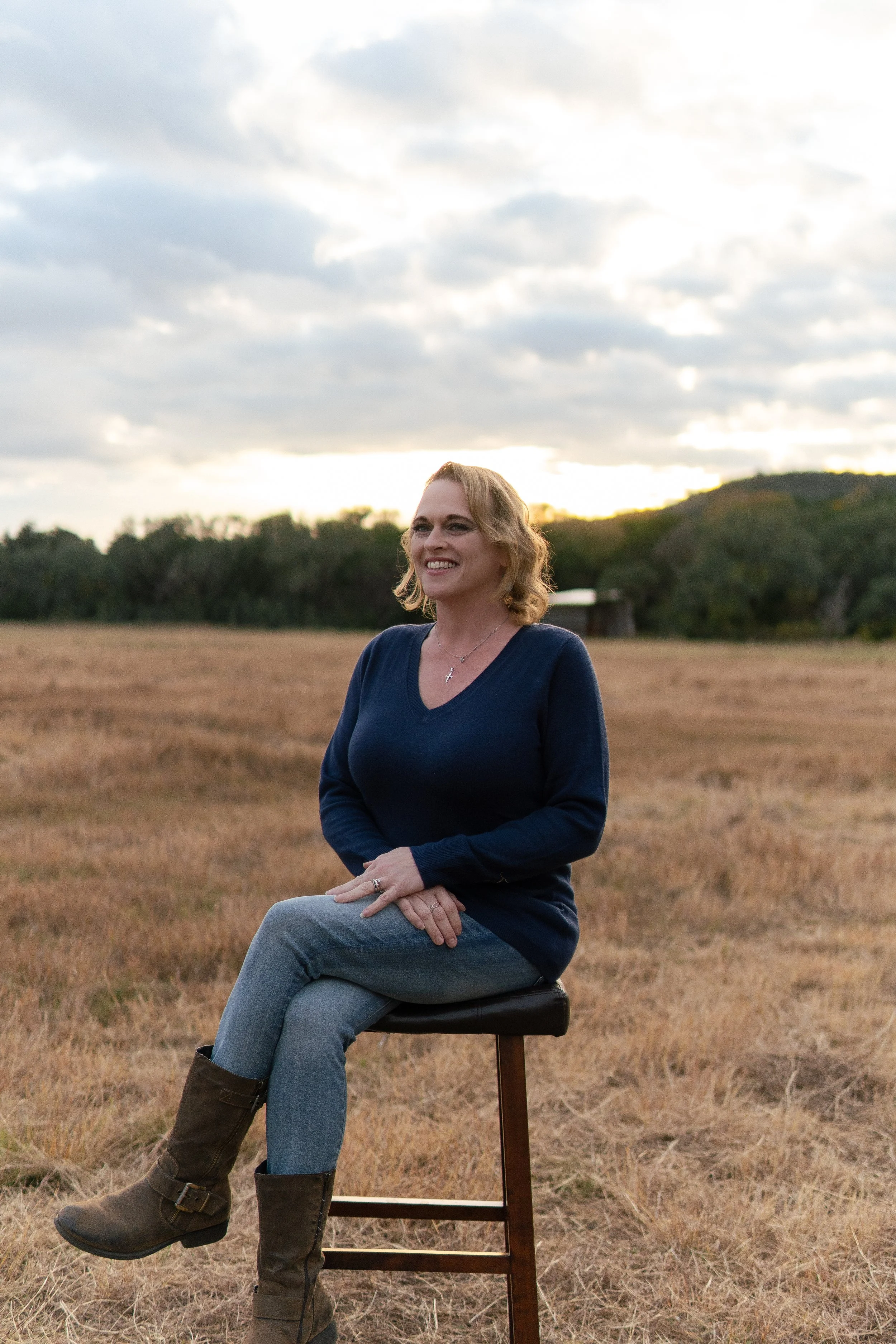 A woman sitting on a chair in a field with a background of hills and cloudy sky during sunset or sunrise.