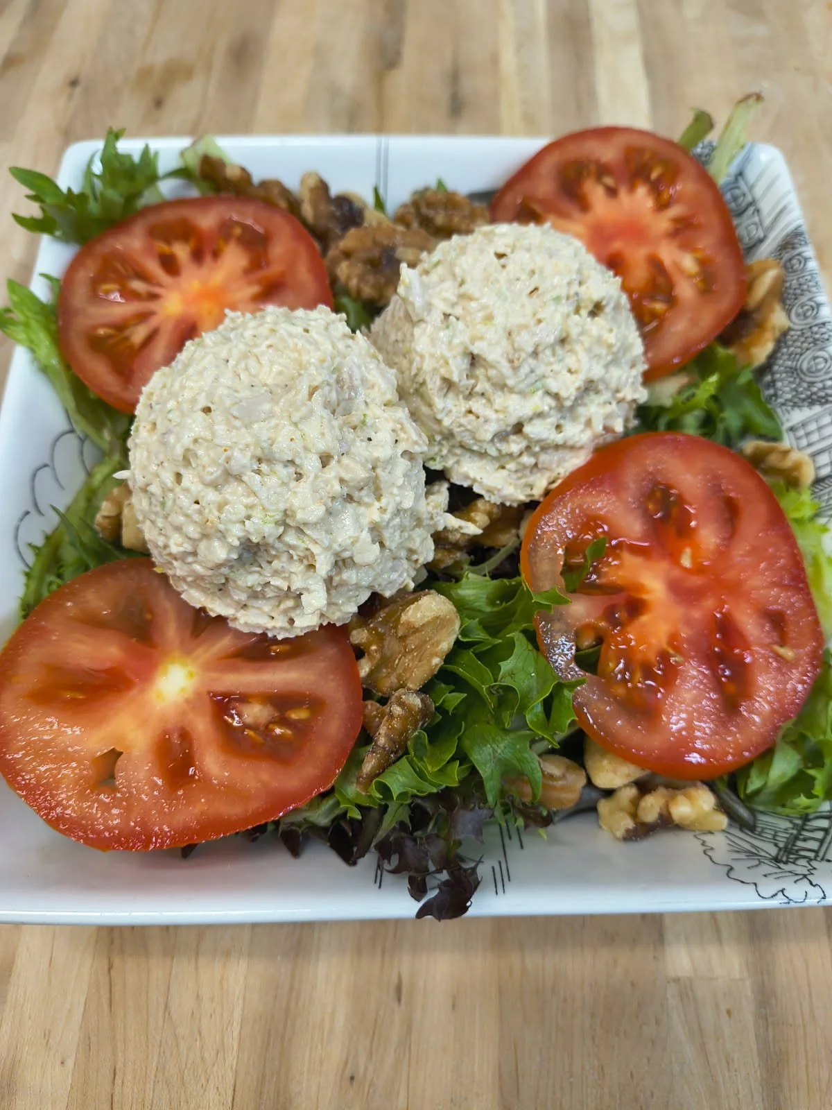 Salad with sliced tomatoes, walnut pieces, and two scoopfuls of chicken or tuna salad on leafy greens in a decorative bowl.