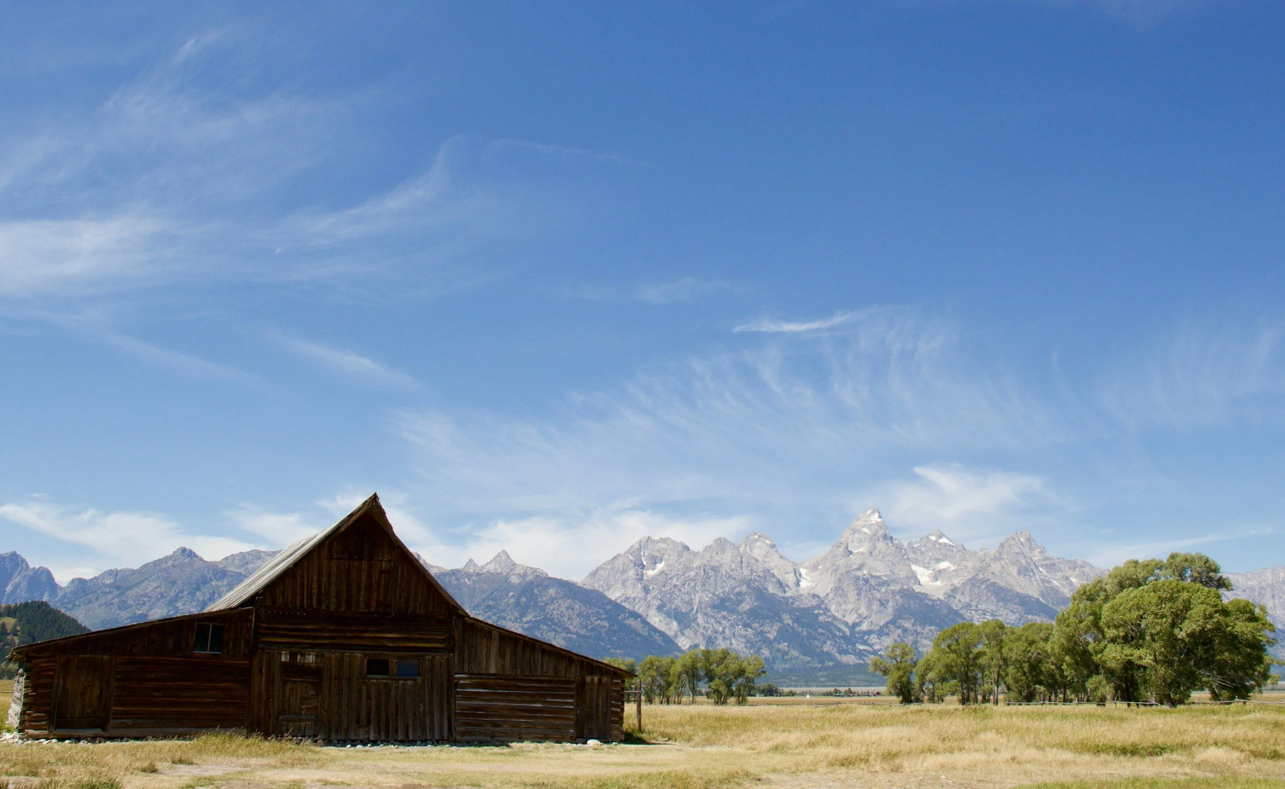 A rustic wooden barn in a field with mountains in the background under a blue sky with wispy clouds. The overall farm support dream.