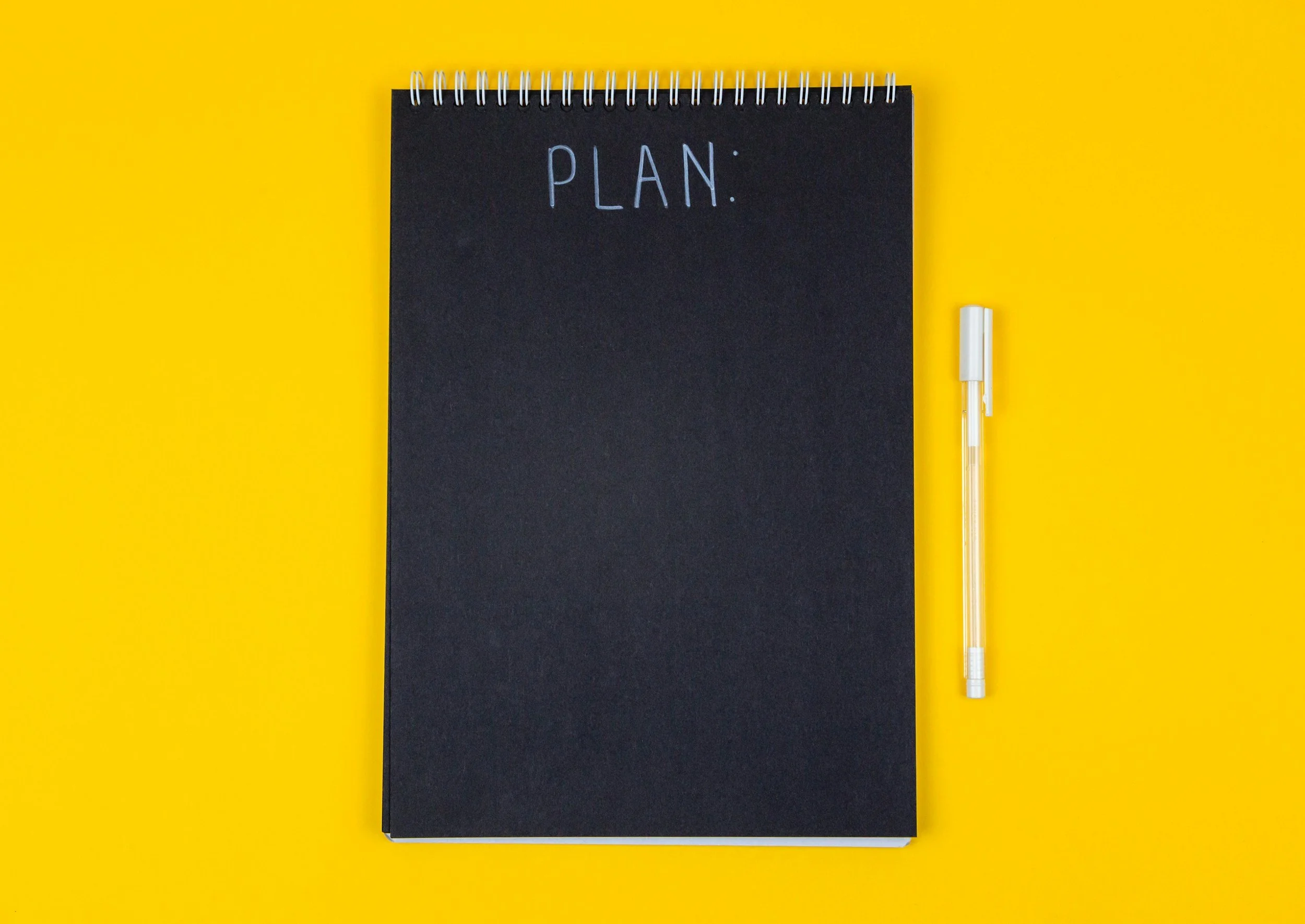 Black spiral notebook with the word ''PLAN'' written on the cover and a yellow background, with a white pen beside it. Representation of a grant funding and strategy plan.