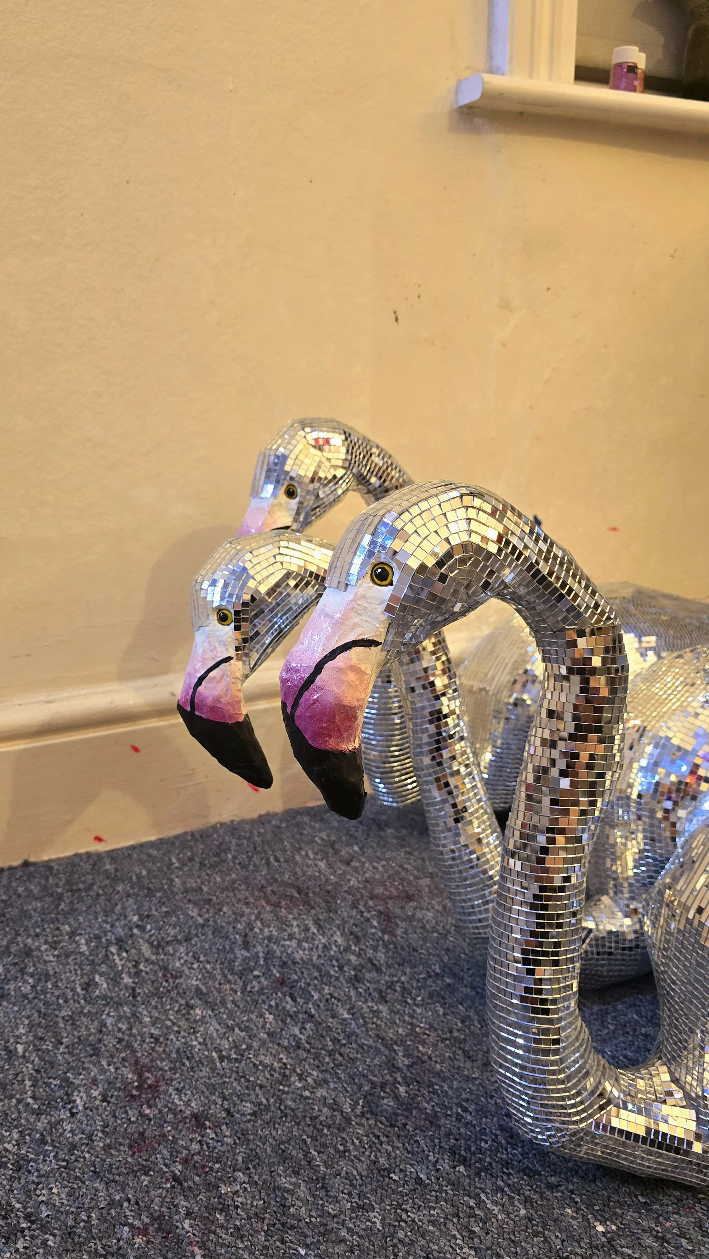 Three decorative flamingo sculptures with mirrored mosaic bodies and painted heads, standing on a floor against a beige wall.