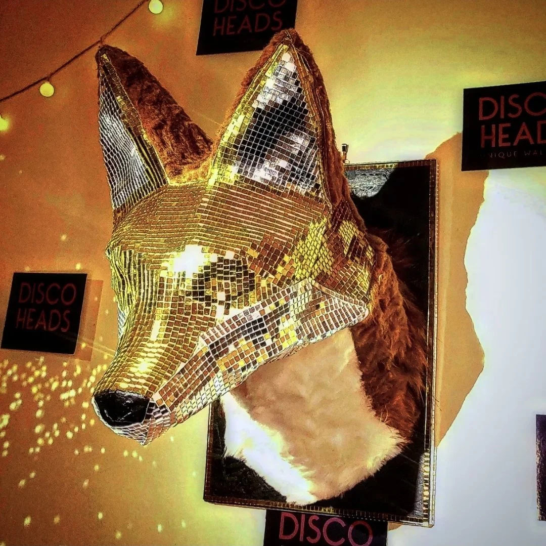 Decorative art piece of a dog's head with a mosaic mirrored surface and real fur on the sides, displayed on a wall with yellow lighting and black signs labeled 'DISCO HEADS'.