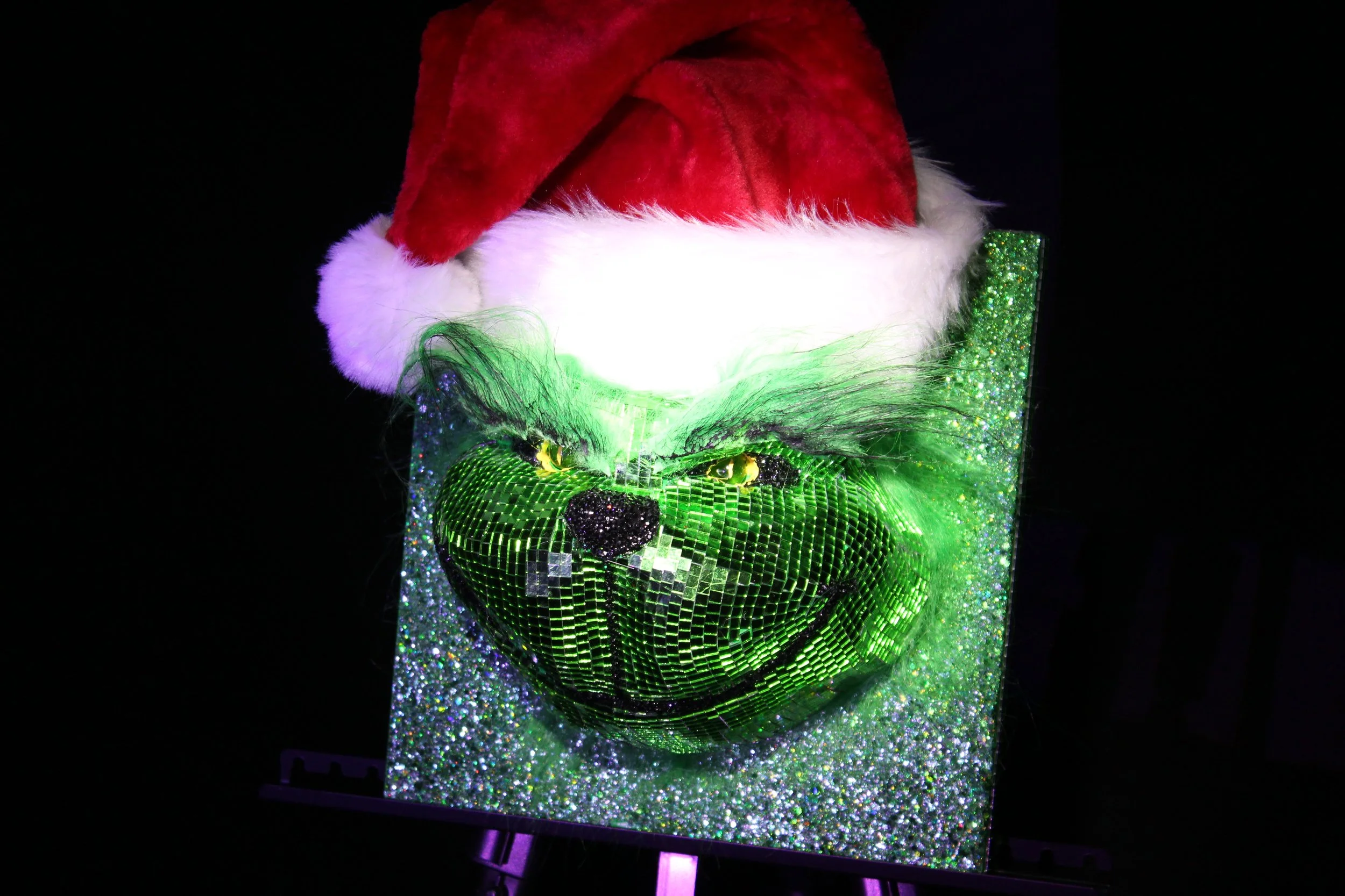 A Santa hat placed on top of a glittering, holographic cube with green and black patterns, illuminated by green and purple light against a black background.
