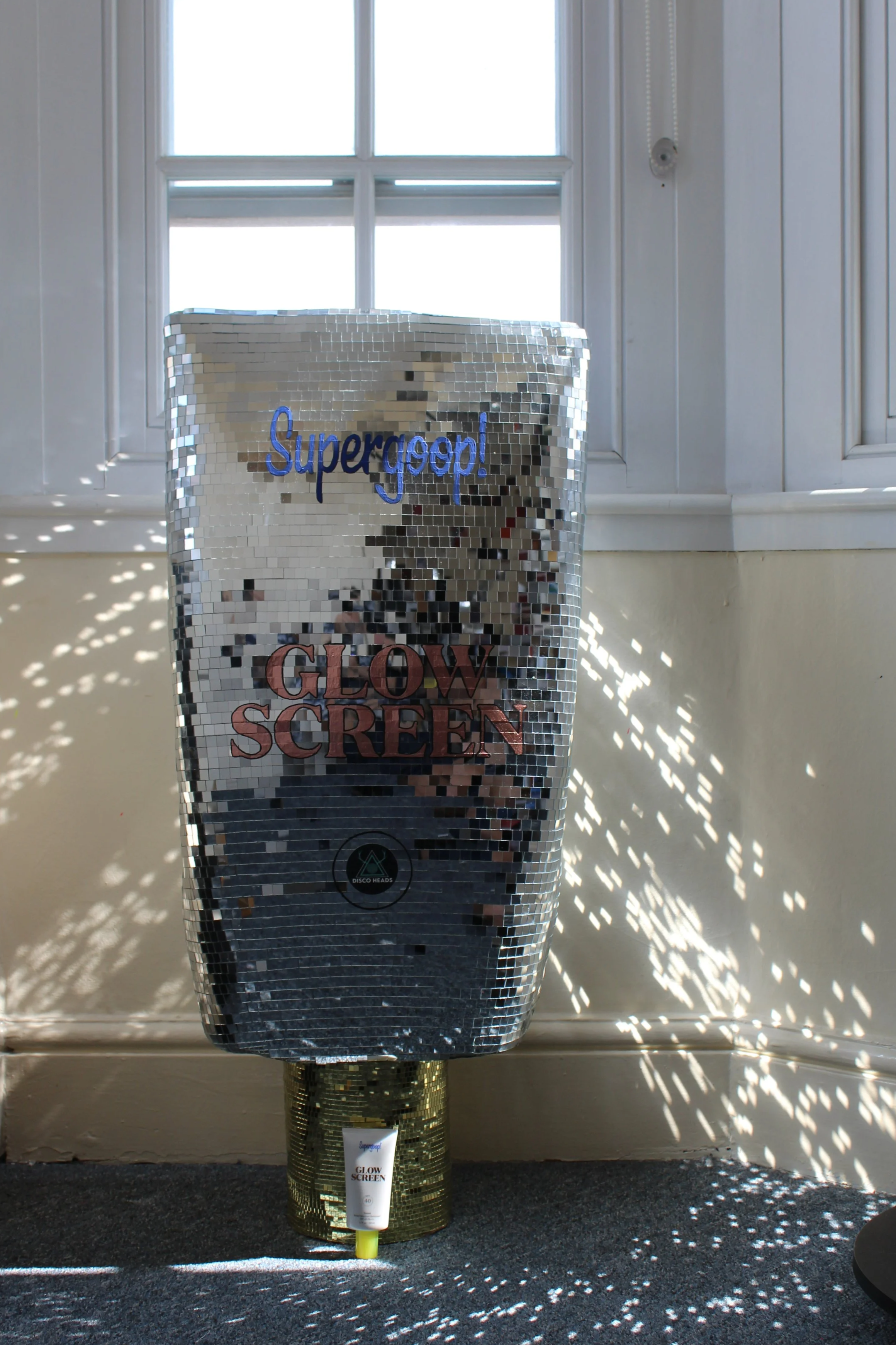 A disco ball style reflective box with the word 'Supergosp!' written on it, placed on a carpeted surface behind a tube of Glow Screen sunscreen. Light and shadow cast patterns on the wall and floor from the reflective box near a window with sunlight.