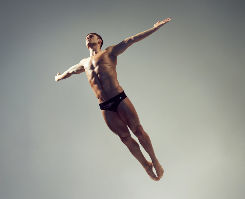 A male gymnast or dancer in mid-air with arms outstretched, wearing black shorts, against a plain background.