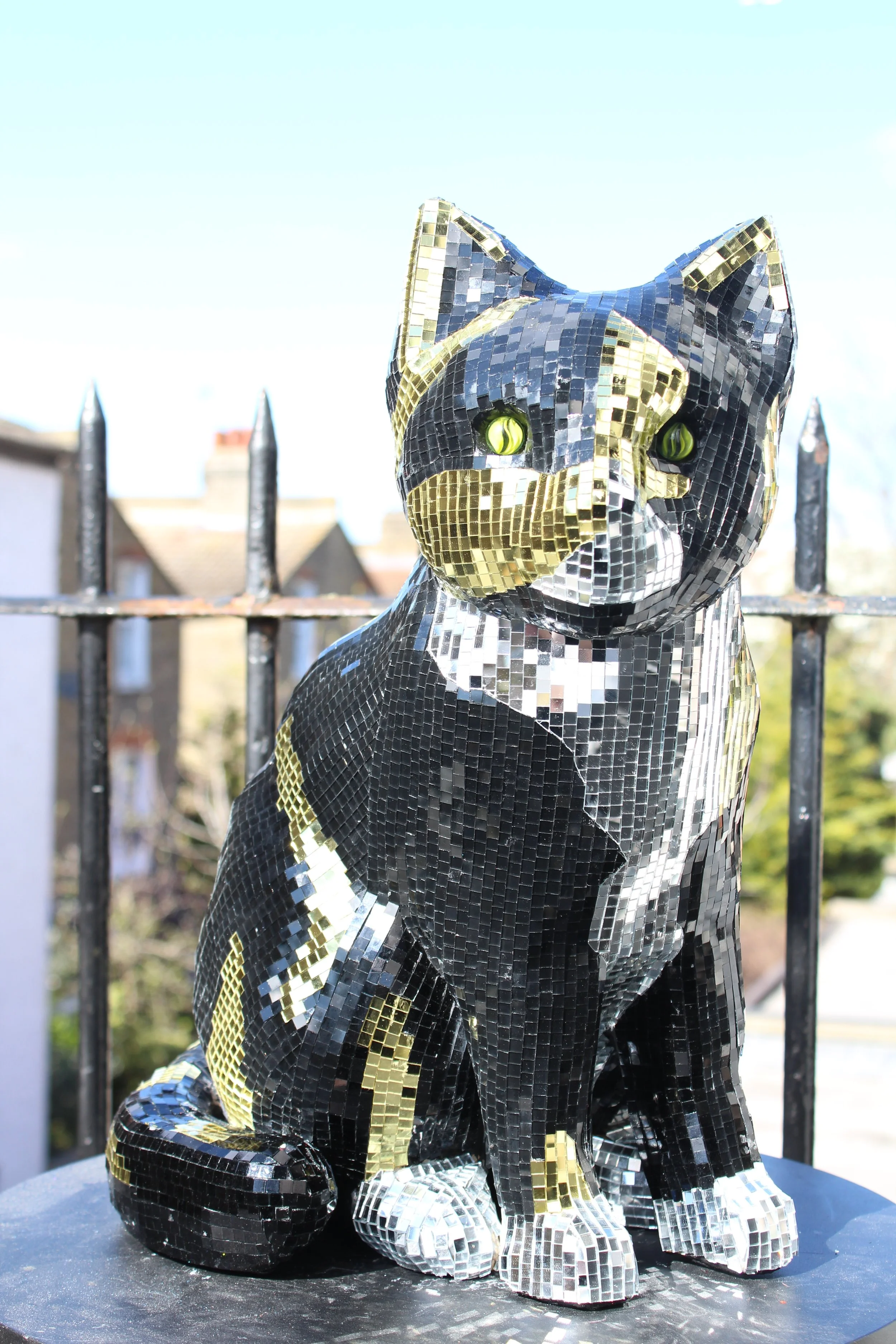 A reflective mosaic sculpture of a sitting dog with black, gold, and silver tiles, situated outdoors behind a metal fence.
