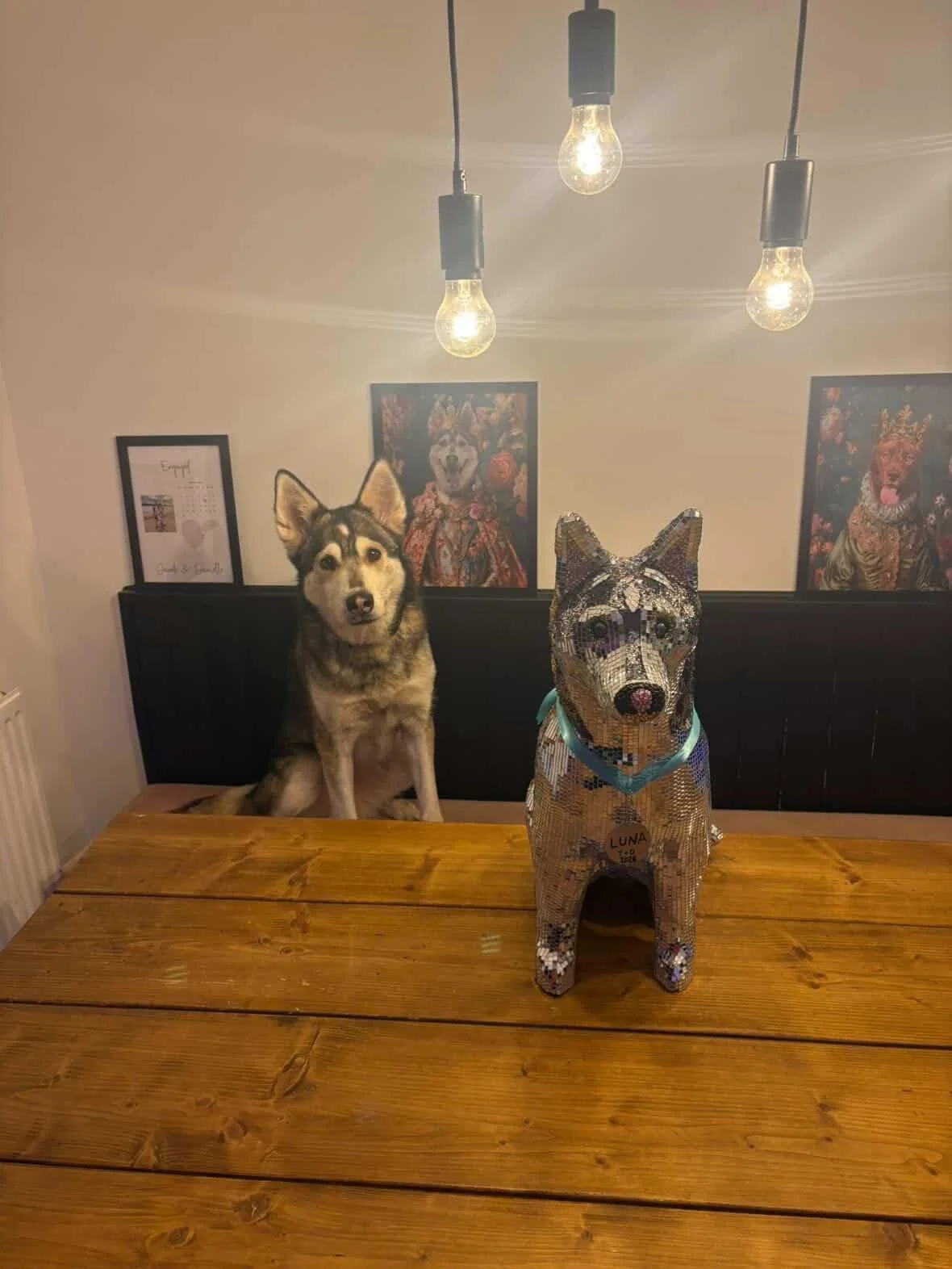 A Siberian Husky sitting at a wooden table next to a mosaic dog sculpture on a wooden table. The background has framed pictures and paintings on the wall, illuminated by three hanging light bulbs.