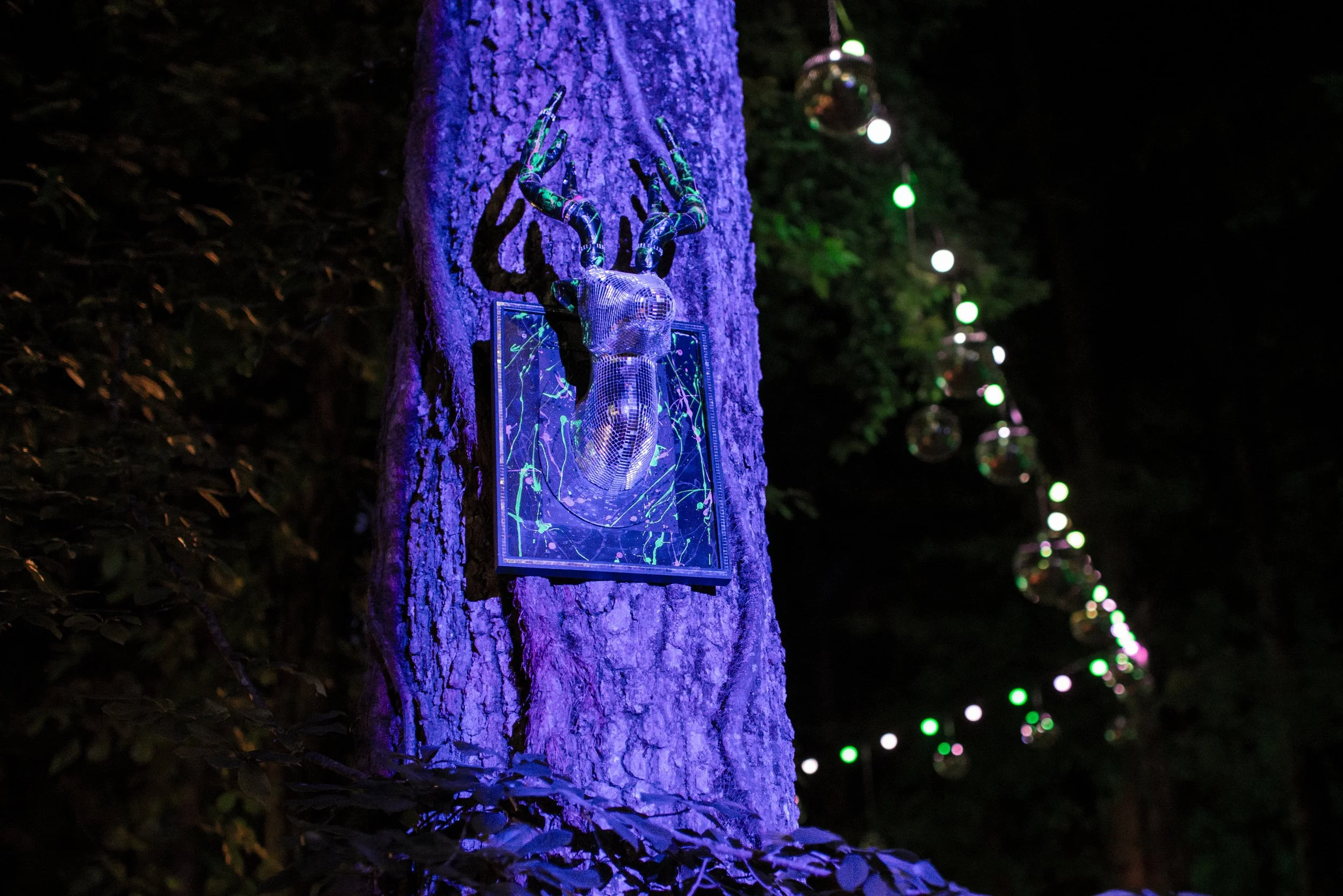 A decorative metallic hand sculpture attached to a tree trunk, illuminated with purple light, with a small framed digital art piece at its base. In the background, colorful string lights hang and a large disco ball reflects light in the dark outdoor setting.