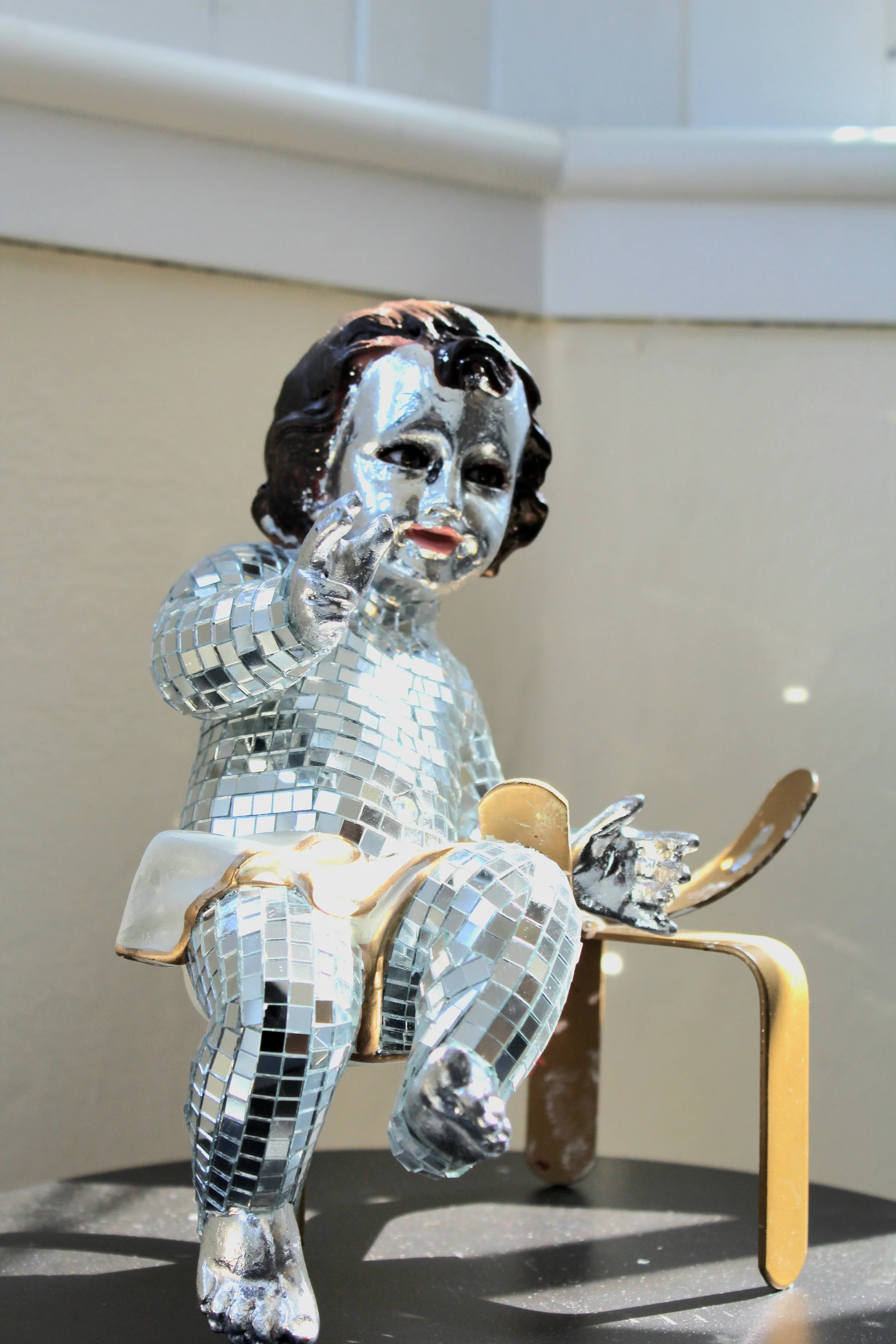 A decorative figurine of a human-like character with a silver face and black hair, dressed in a reflective, mirror-like body suit. The figurine is making a gesture with its hand near its face, with a small golden object wrapped around its wrist, and stands on a gray surface with a blurred background.