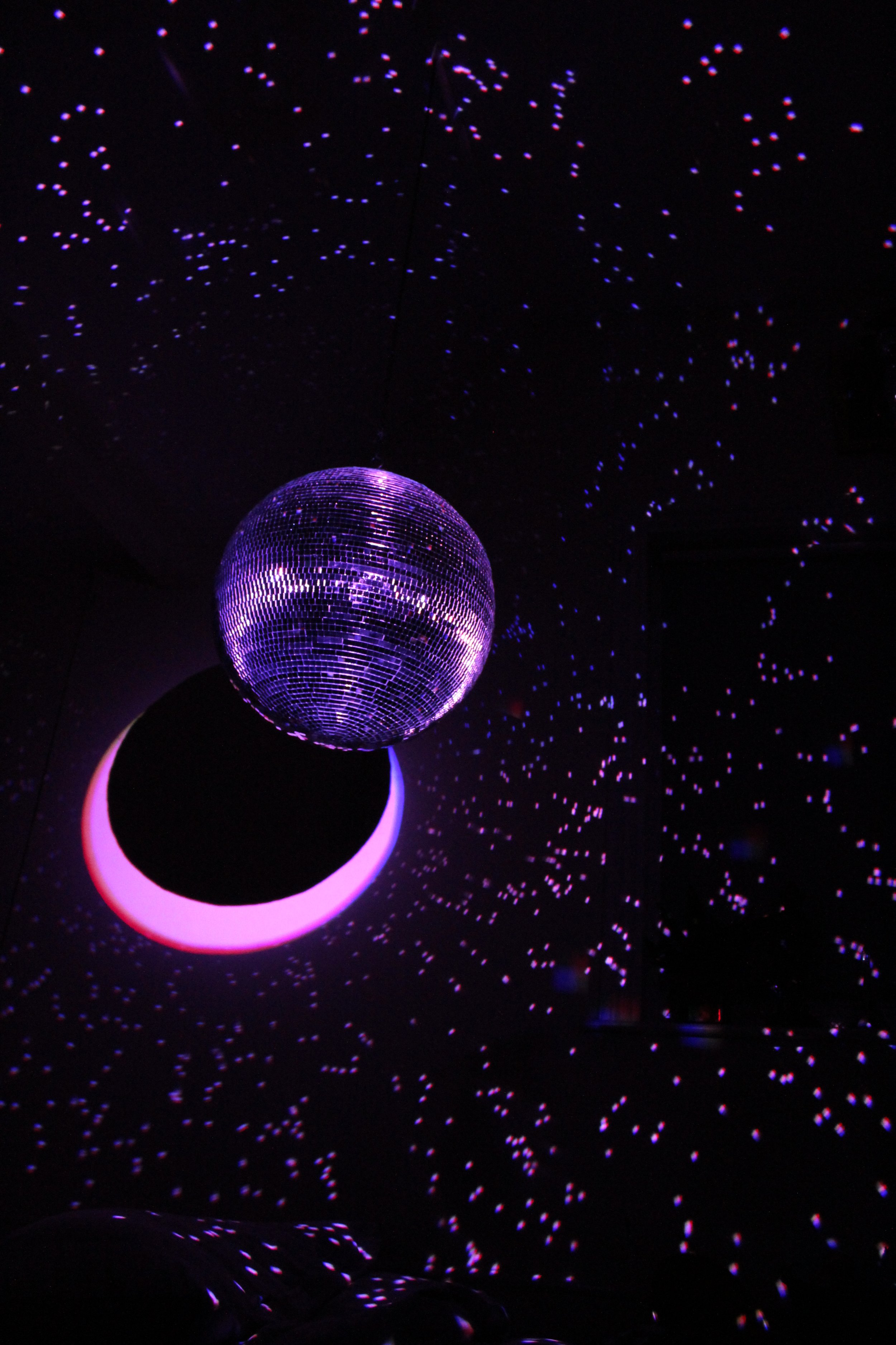 A large disco ball reflecting purple lights hangs from the ceiling. Surrounding it are tiny purple and red lights scattered around, creating a starry effect against a dark background. A neon pink crescent shape is present below the disco ball.