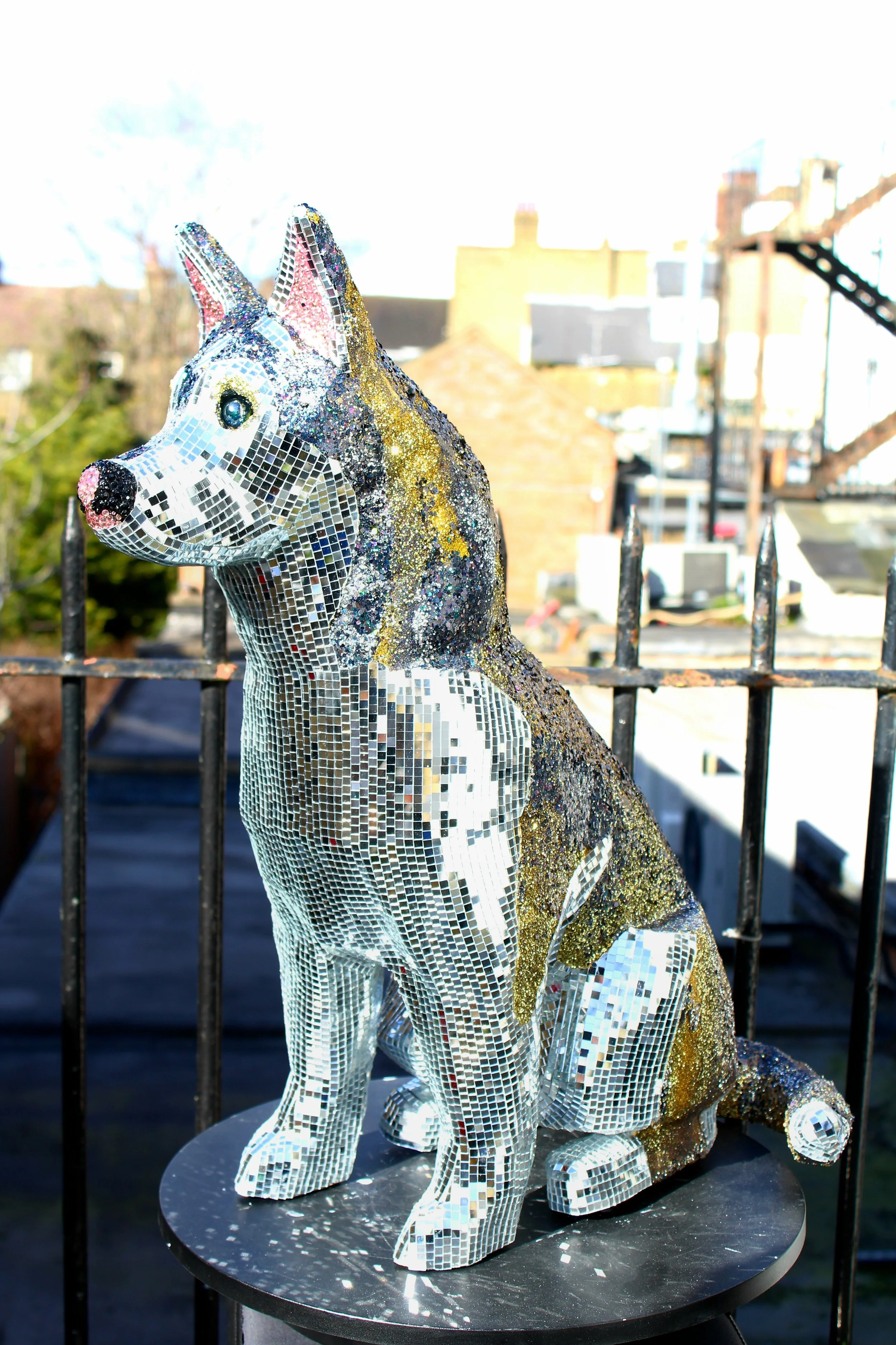 A life-sized mosaic sculpture of a dog sitting on a black circular platform outdoors, surrounded by a metal fence, with buildings and leafless trees in the background.
