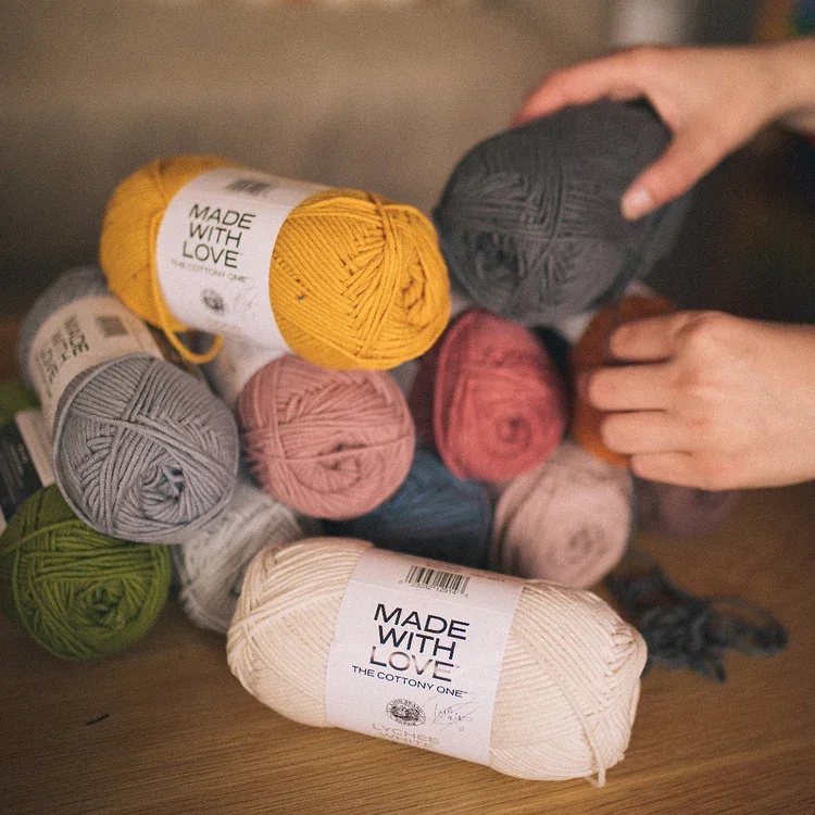 Multiple skeins of yarn in various colors, including yellow, gray, pink, red, green, cream, and navy, are arranged on a wooden surface. Two hands are reaching to pick up or adjust the yarn. The yarn skeins have labels with the text "Made With Love".