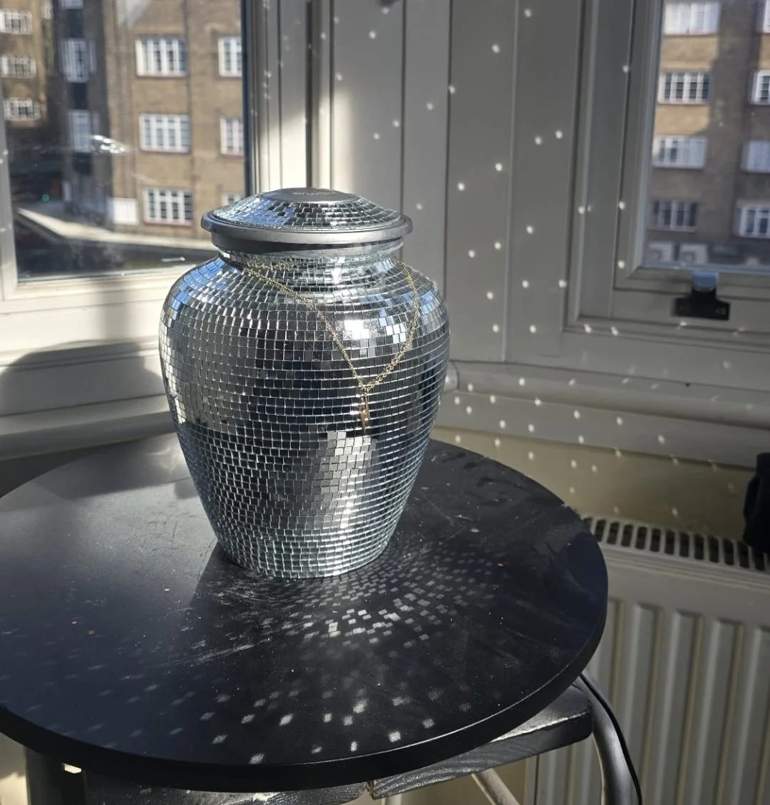 A small silver disco ball decorated with a gold chain and pendant sitting on a black tabletop in front of a window, with sunlight creating a pattern of light spots on the wall and table.