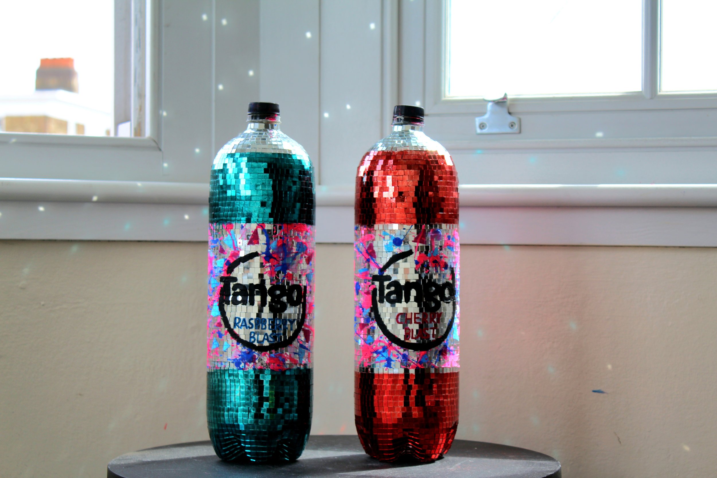 Two bottles of Tanggo soda, one lime green and one cherry red, covered in a mosaic style reflective surface, placed on a dark pedestal by a window with white framing.