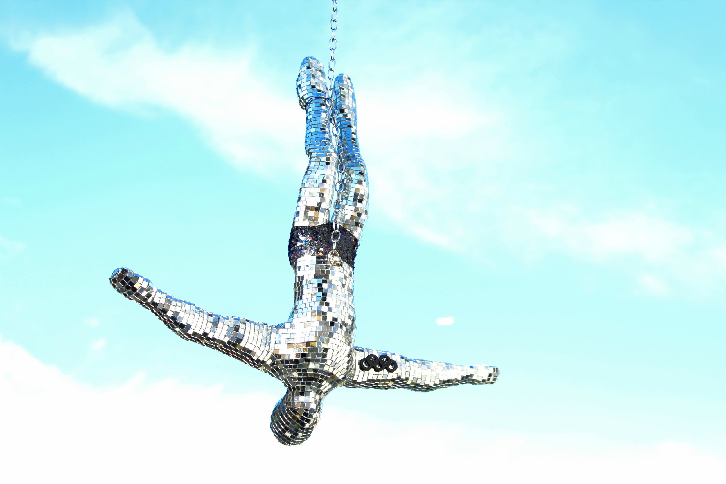 A human figure made of small reflective tiles in a yoga pose against a blue sky, with the Olympic rings symbol on the wrist.
