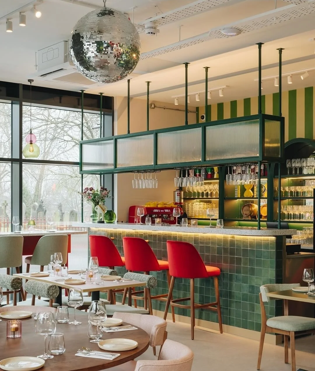 Modern restaurant interior with large window, disco ball, green tile bar with red bar stools, hanging glasses, and a variety of dining tables with chairs and glassware.