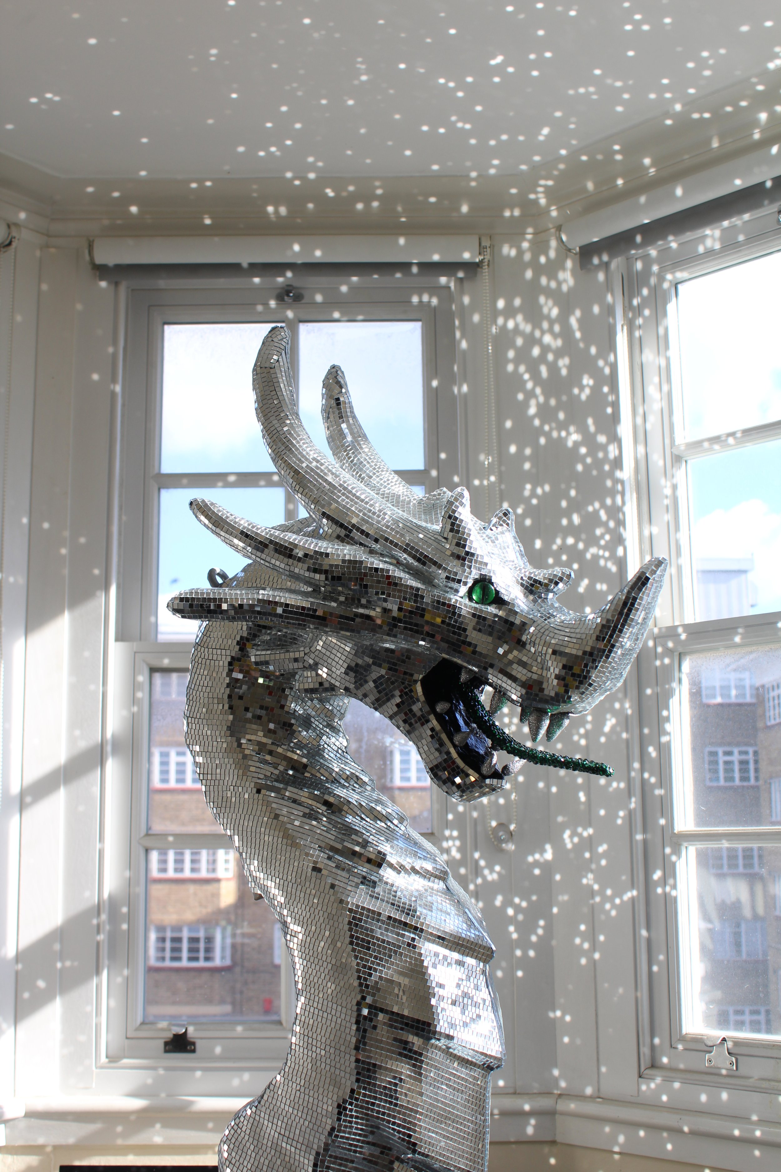A mirror sculpture of a dragon's head and neck made of small, reflective tiles, positioned in front of a window. Sunlight through the window creates a pattern of small light spots on the wall and ceiling.