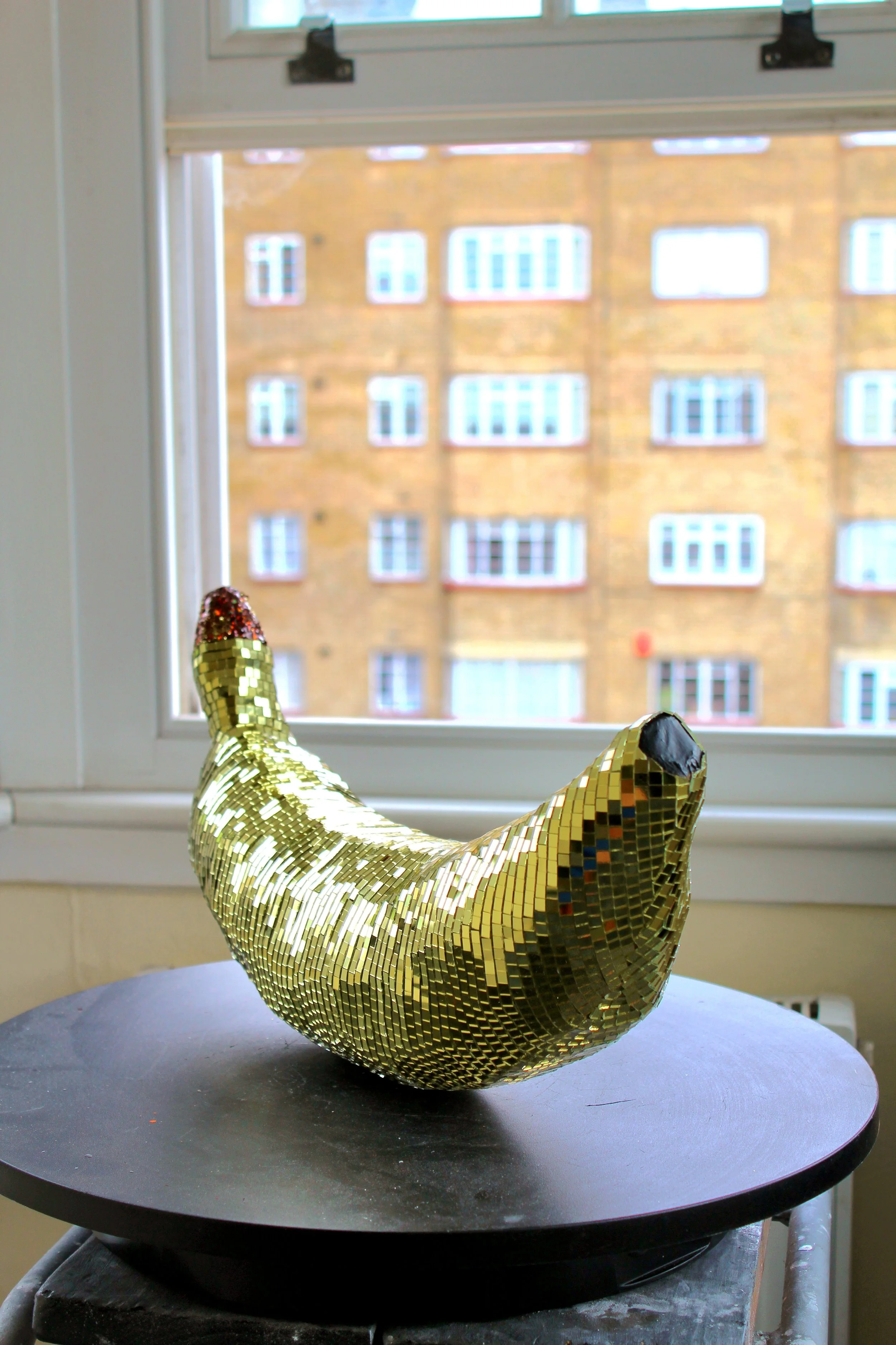 A pair of shiny, gold, mosaic-style, high-heeled shoes on a black circular table with a window and building in the background.