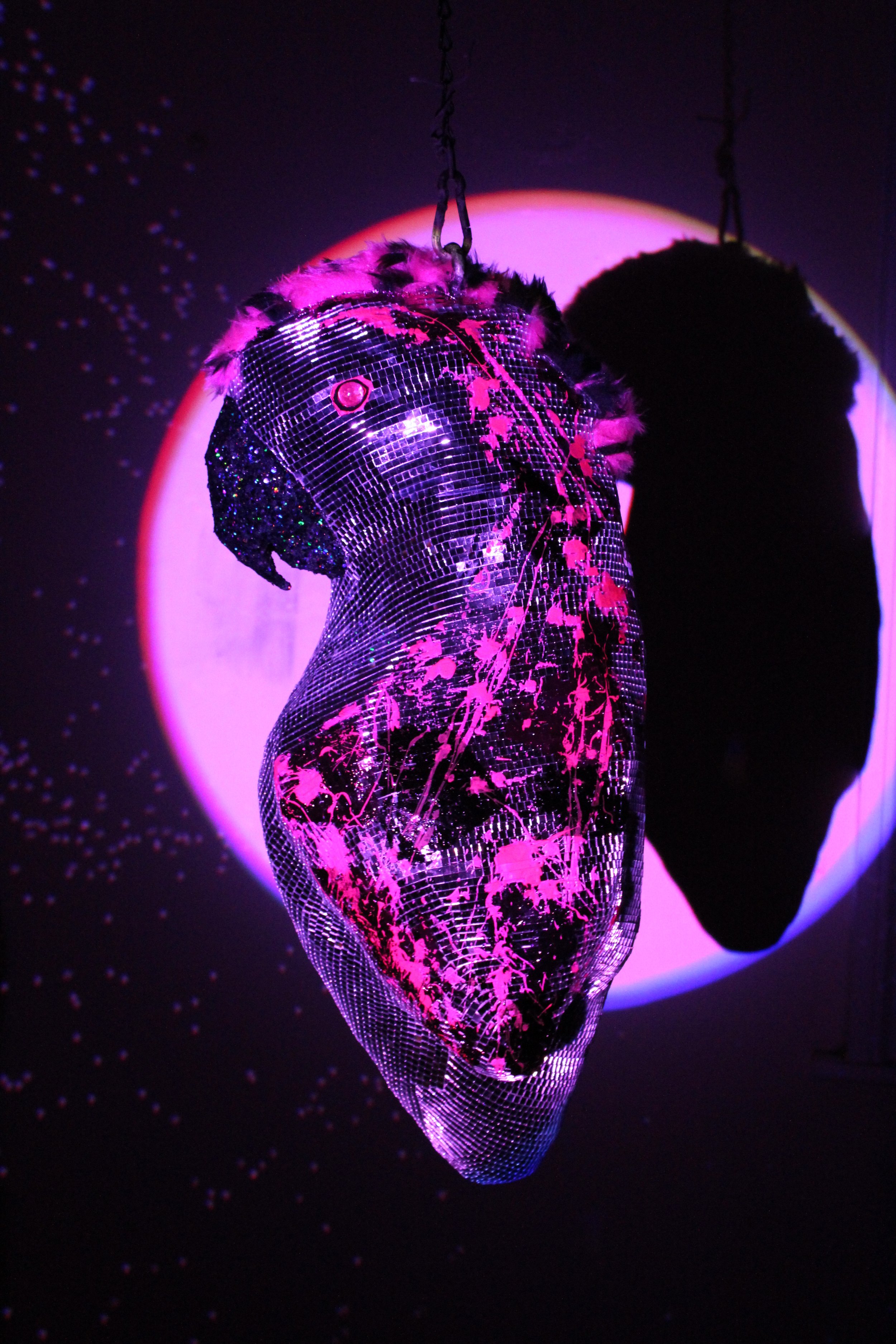 A shiny, reflective disco ball with pink paint splatters hanging from chains in front of a purple-lit circular backdrop.