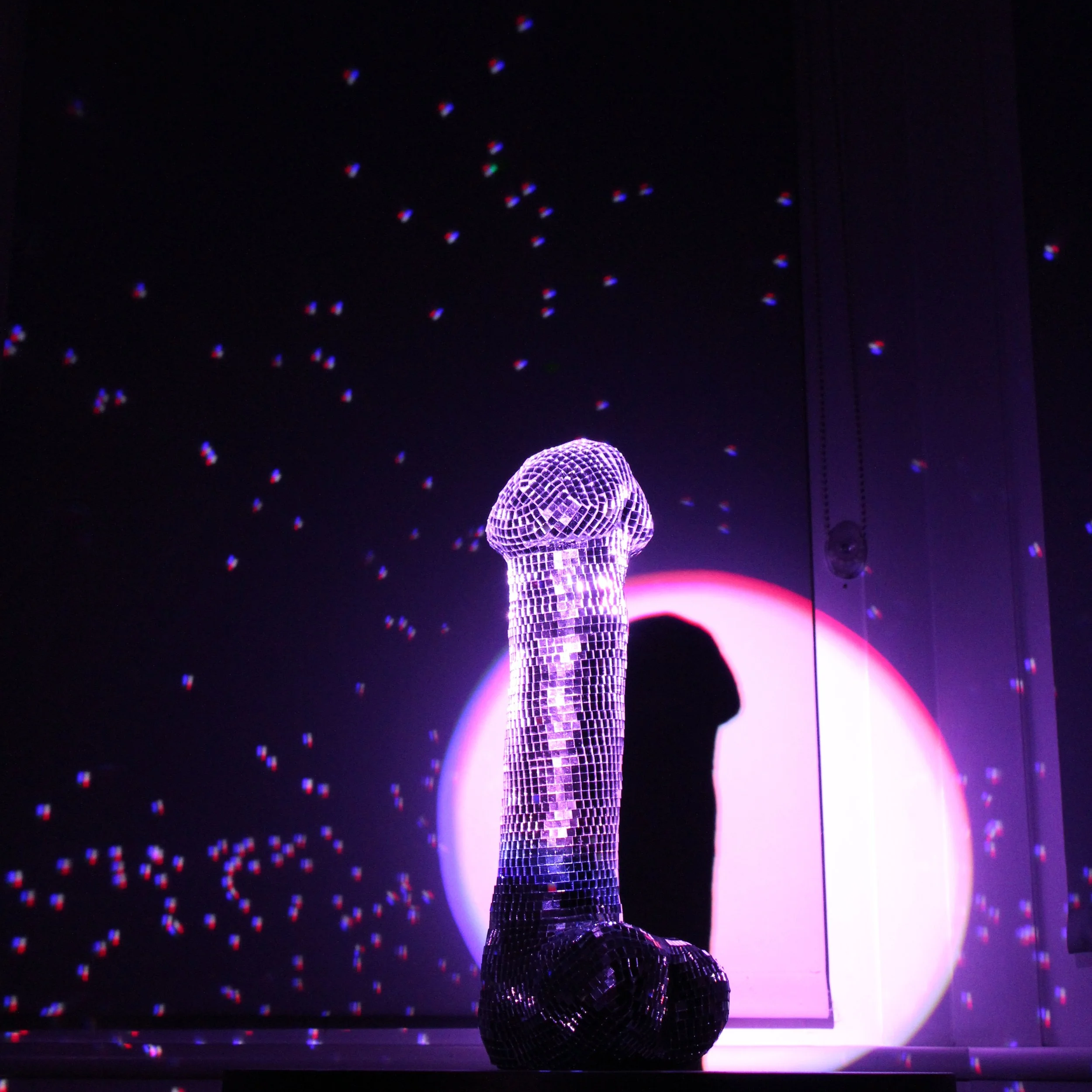 A low-lit room with a disco ball on the ceiling and a figure covered in small reflective tiles, illuminated by purple and reddish lights.