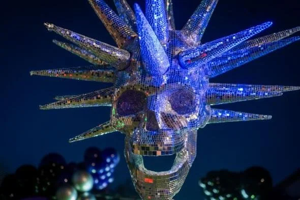 A shiny, reflective sculpture of a face with spiky, hair-like projections made of mirrored tiles, illuminated with blue and multicolored lights.