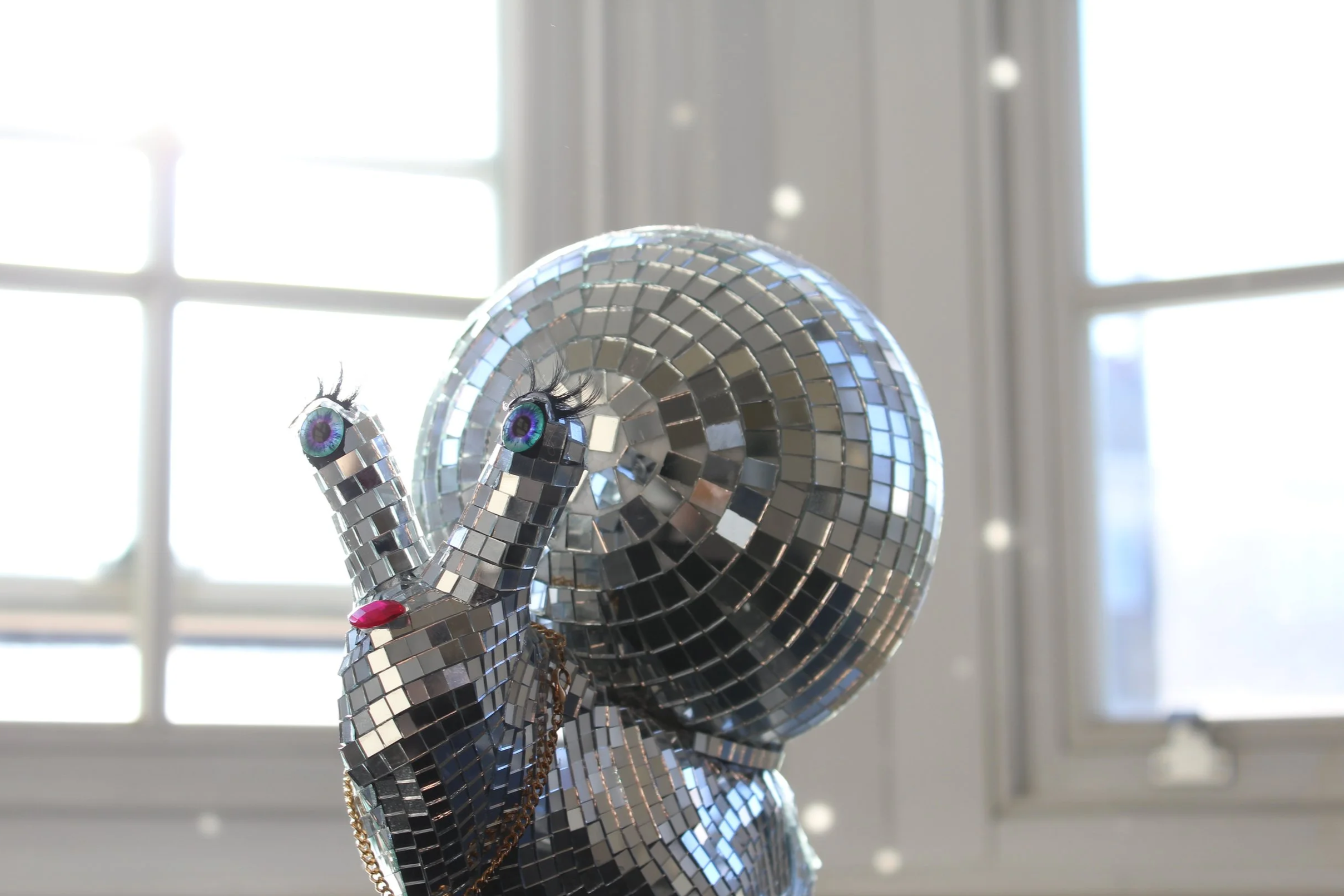 A decorative sculpture resembling a snail with a spiral shell made of small, reflective tiles, featuring large, colorful eyes with long eyelashes and a pink tongue, set against a bright window background.