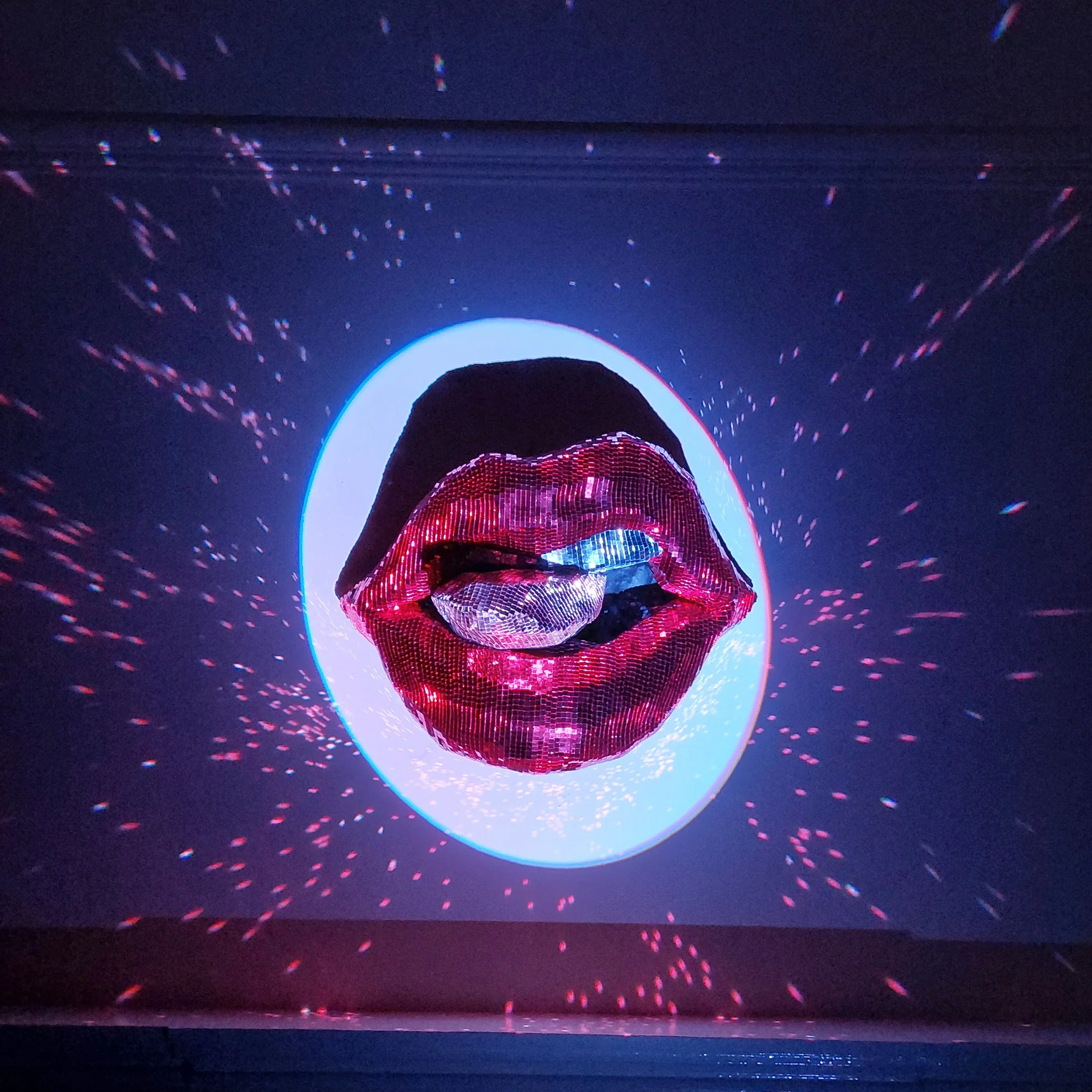 A person wearing a digital mosaic face mask stands in front of an illuminated halo with rainbow-colored light effects against a dark background.