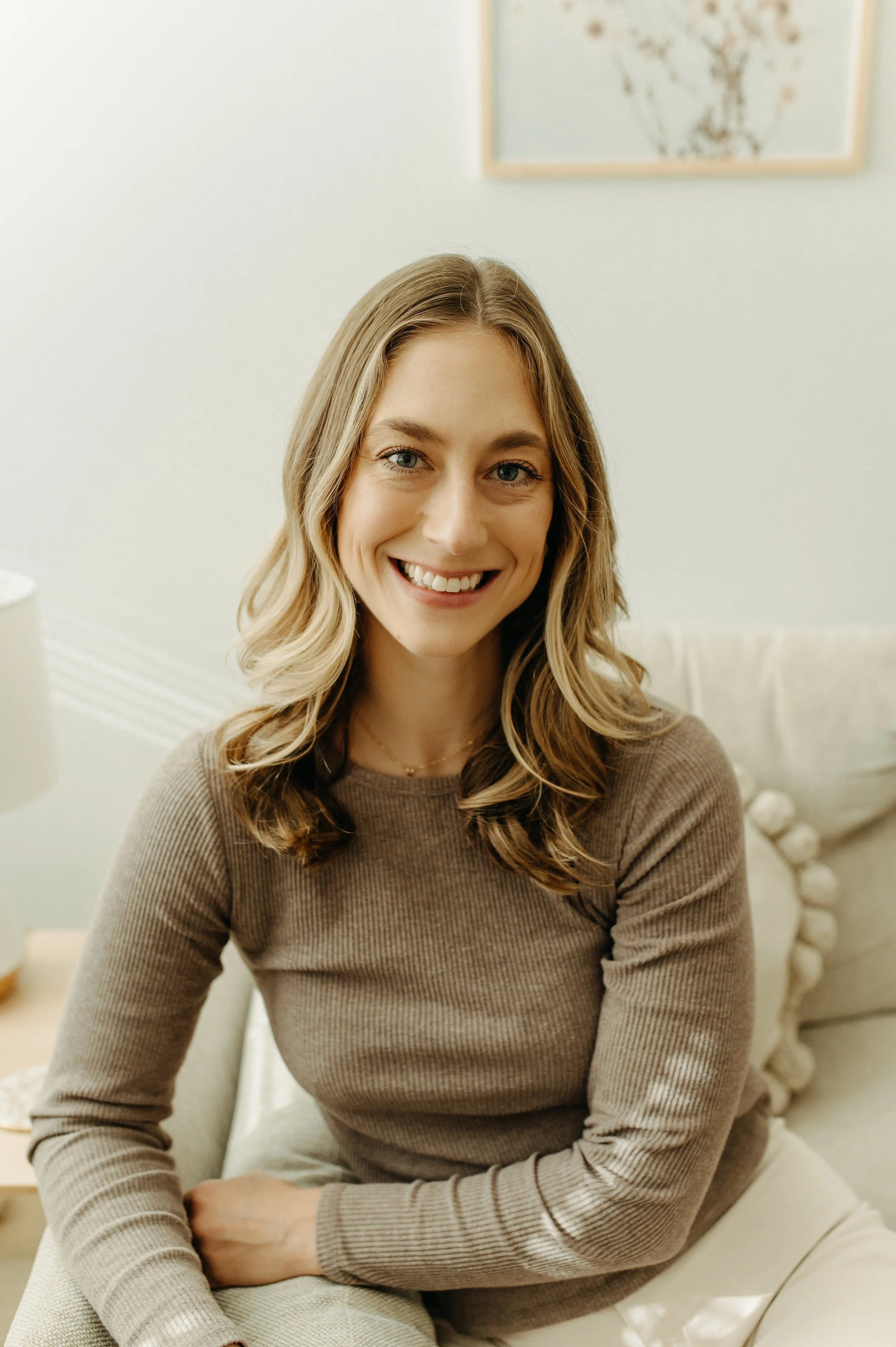 A woman with wavy blonde hair, blue eyes, and a bright smile sitting on a cushioned chair in a cozy, well-lit room with white walls and a framed artwork of a deer head in the background.