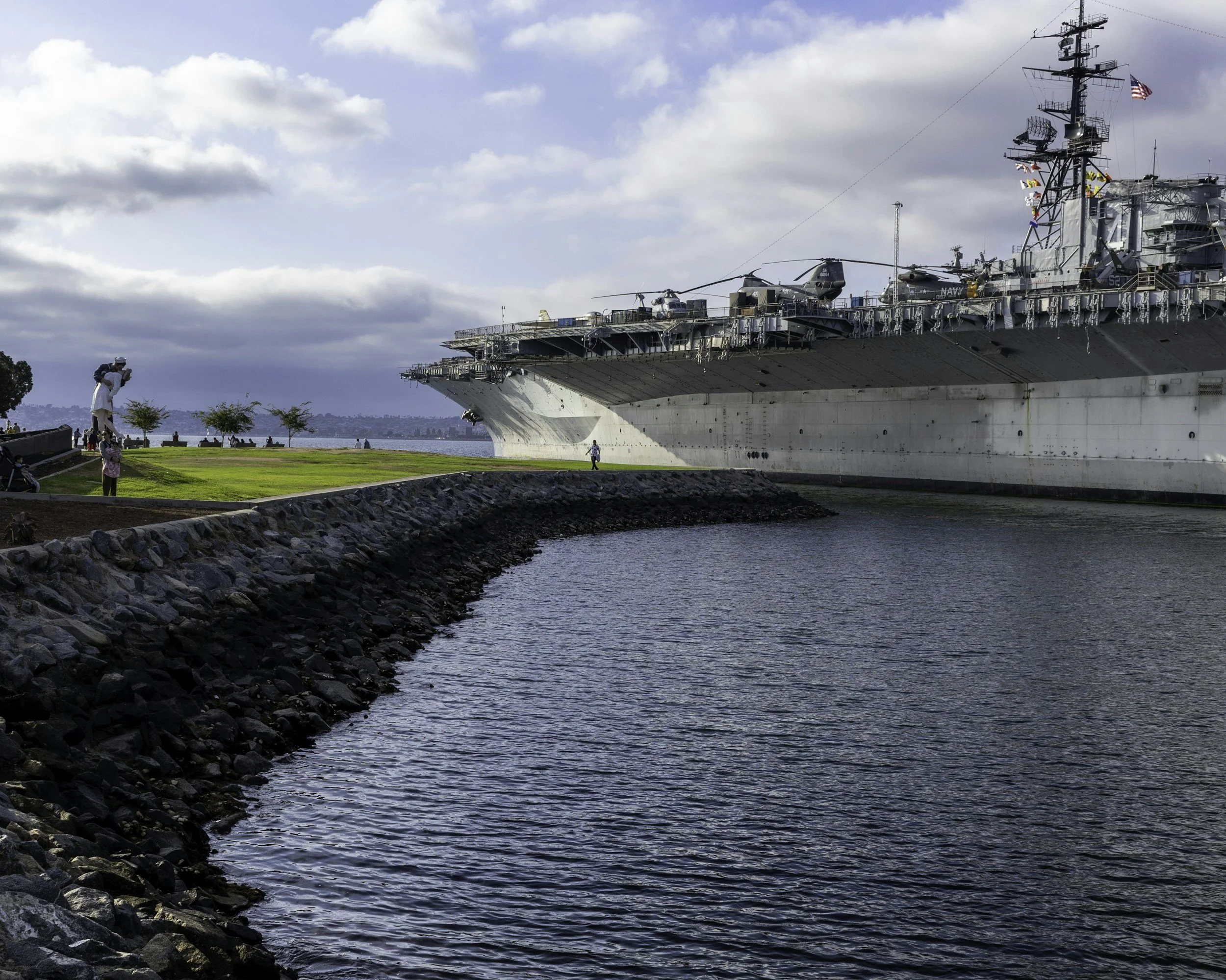 The image shows a close-up view of a large naval aircraft carrier docked at a harbor with people taking photographs nearby. The sky is partly cloudy.