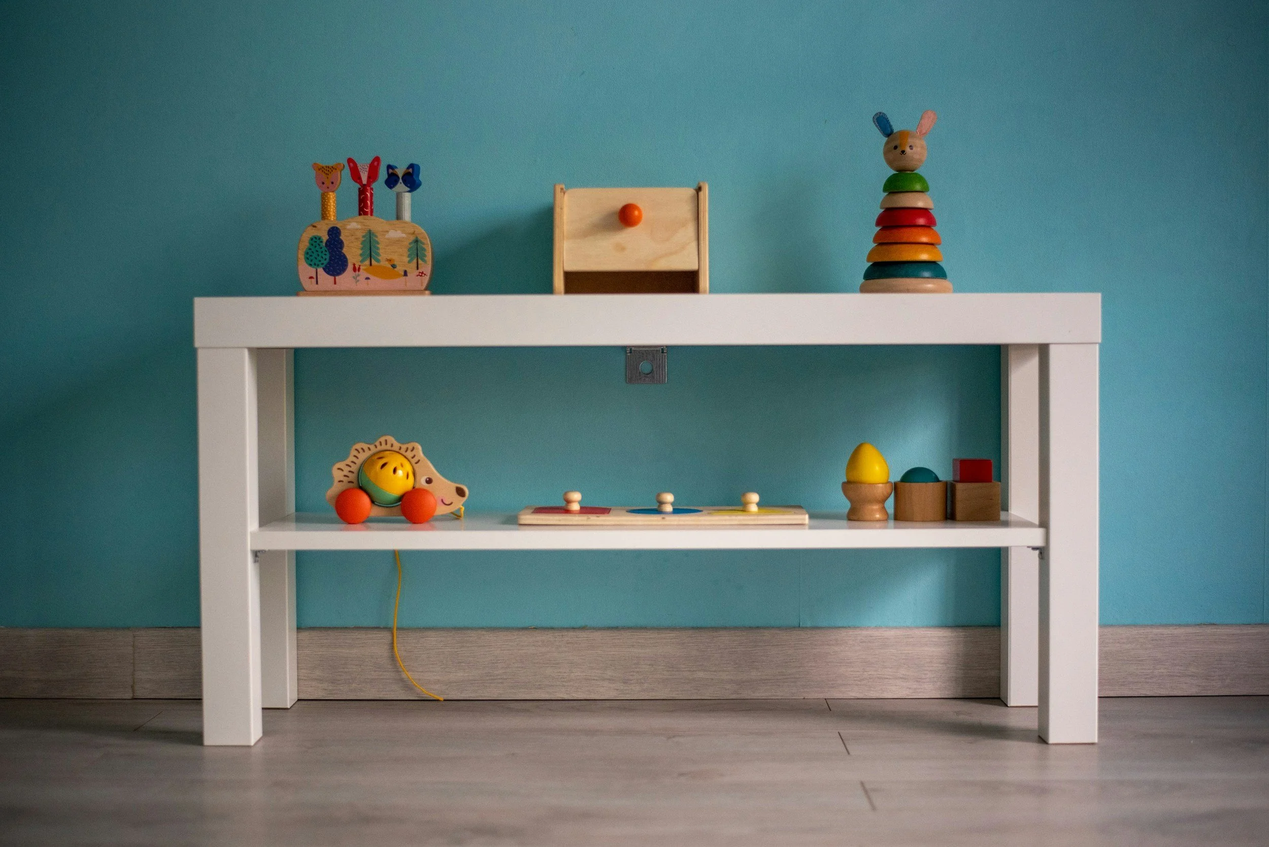 Children's wooden toys arranged on a white shelf against a turquoise wall, including animal figures, stacking rings, a puzzle, and a small wooden box.