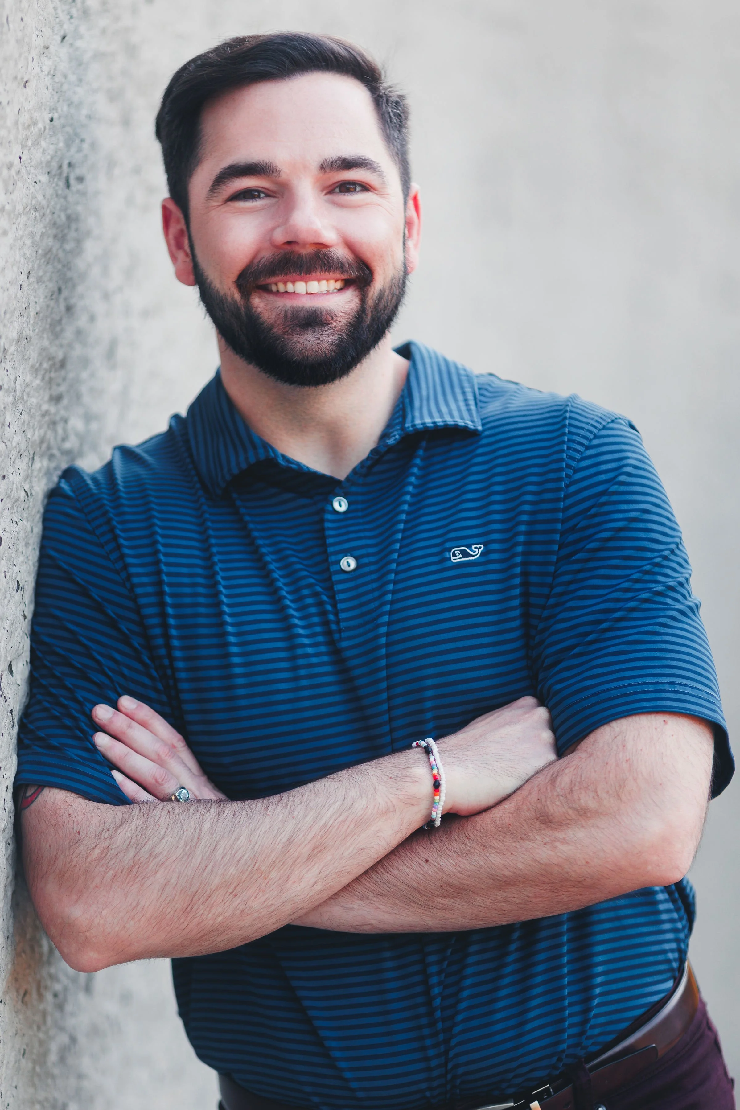 A smiling man with dark hair and a beard standing with arms crossed against a light-colored wall, wearing a blue striped short-sleeve shirt and colorful bracelets.