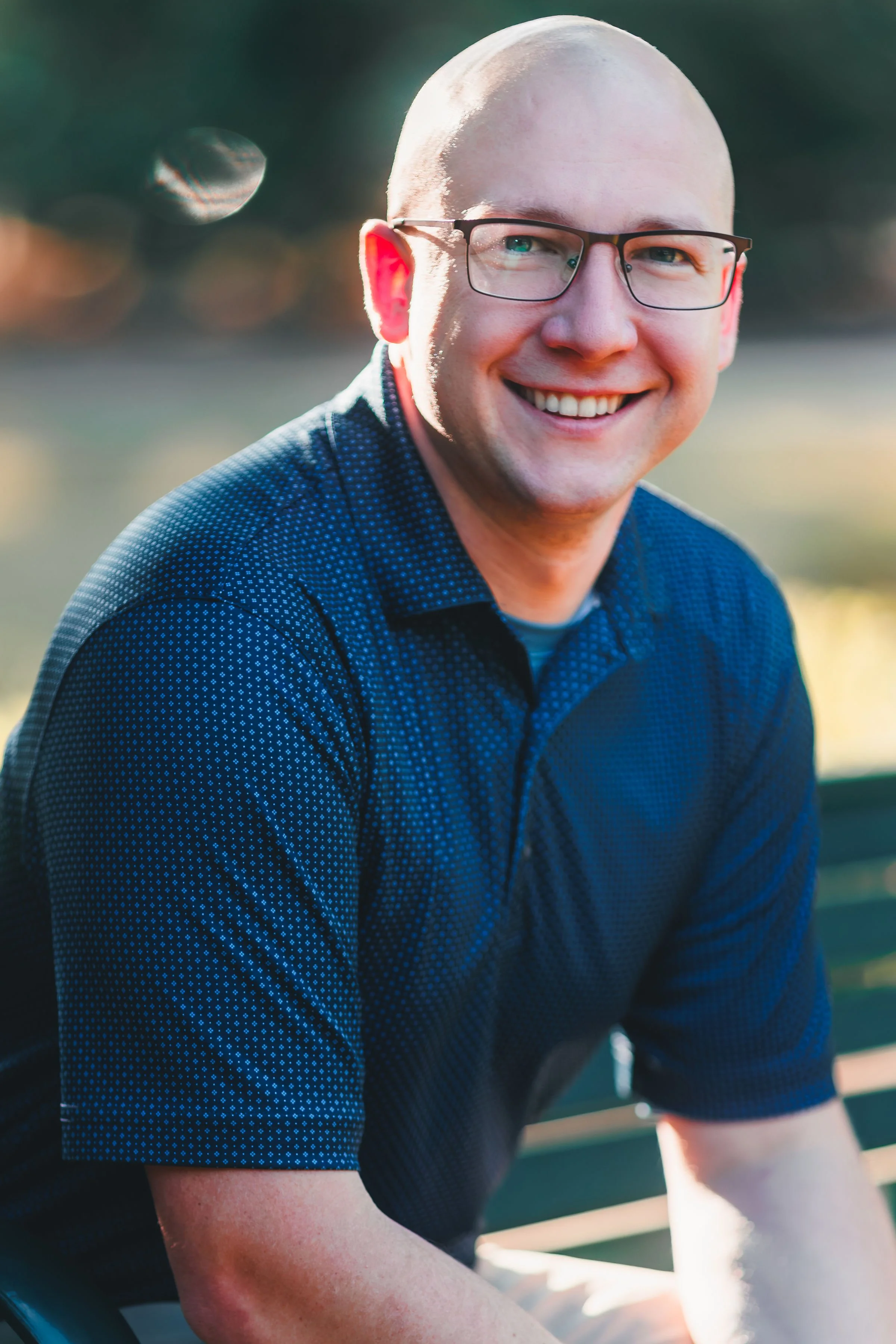Jeremy Garlough, A smiling man with glasses, a bald head, wearing a blue patterned short-sleeve shirt, sitting outdoors during sunset or late afternoon.