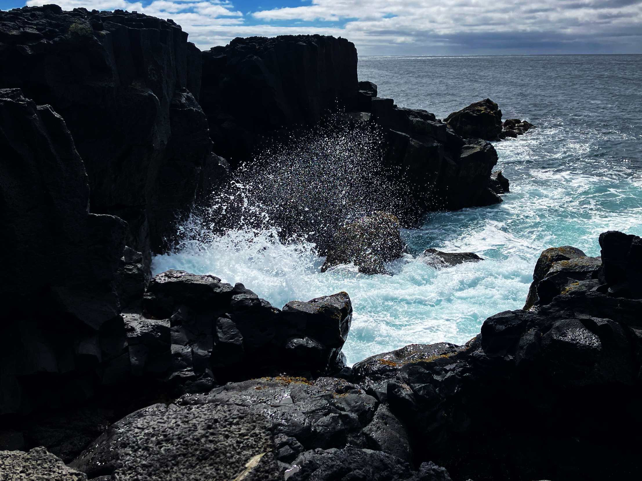 Rocky coastline with black volcanic rocks and ocean waves crashing against them, partially cloudy sky in the background.