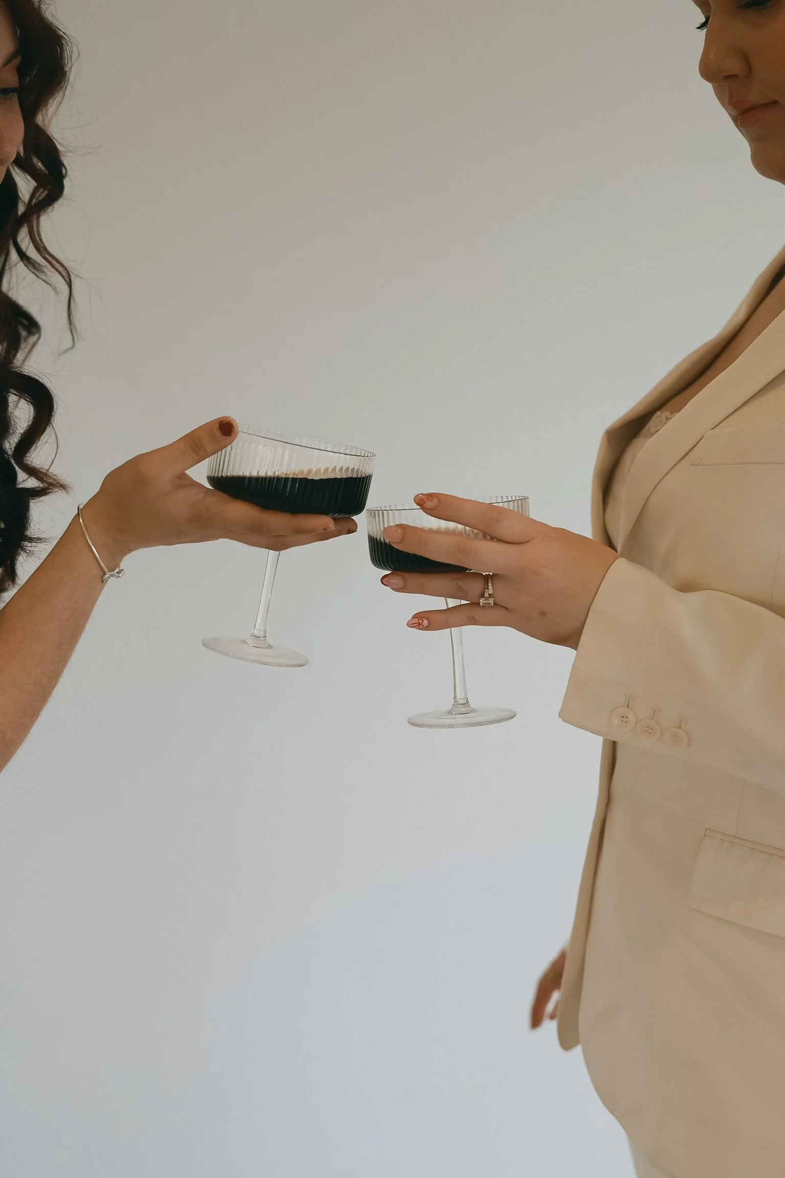 Two women exchanging drinks, one wearing a beige blazer and the other with dark hair, holding glasses filled with dark beverage, against a plain white background.