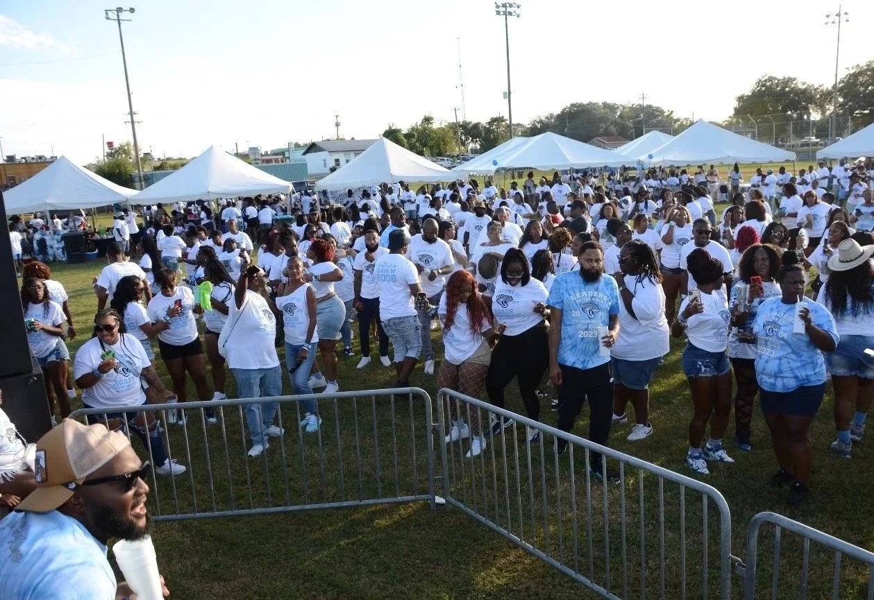 A large outdoor gathering with many people wearing white T-shirts, some with blue designs, at an event with white tents set up on a grassy field. The crowd is standing and socializing, with some people taking photos.