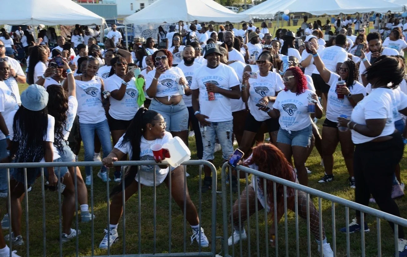 Group of people gathered outdoors at an event, many wearing white t-shirts, some taking photos or videos, with tents in the background.