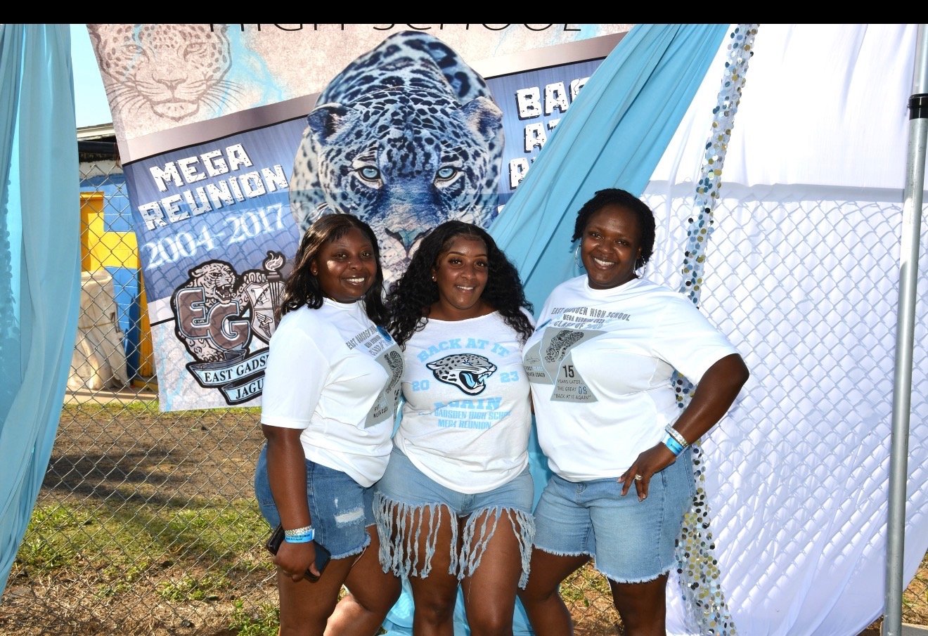Three women standing outdoors in front of a banner with a jaguar, smiling and posing for a photo. They are wearing white T-shirts with a jaguar graphic.