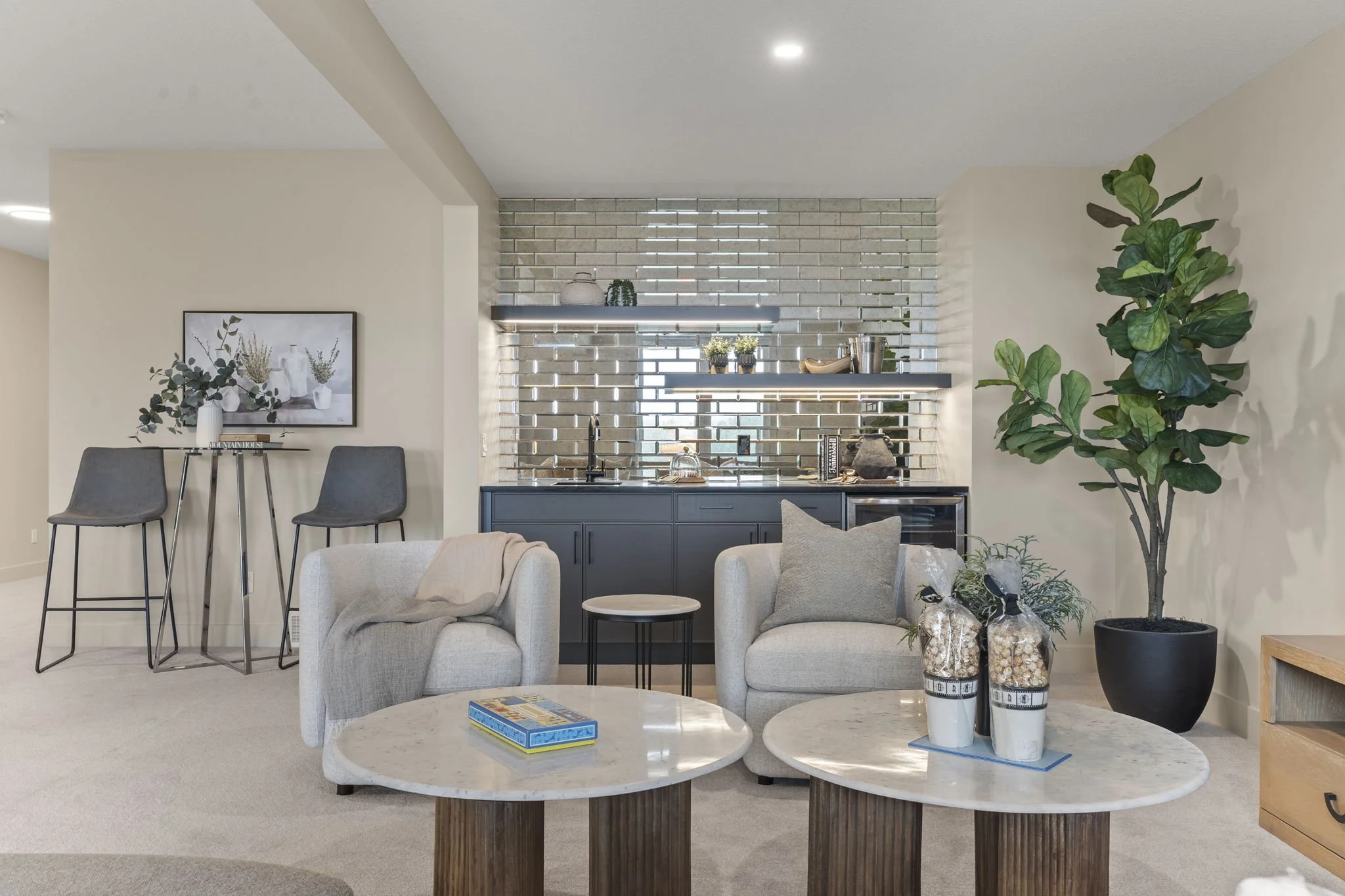 Living room with two beige armchairs and a coffee table with a board game, a large potted plant, a kitchen area with blue cabinets, open shelves, decorative items, and a gray sectional sofa.