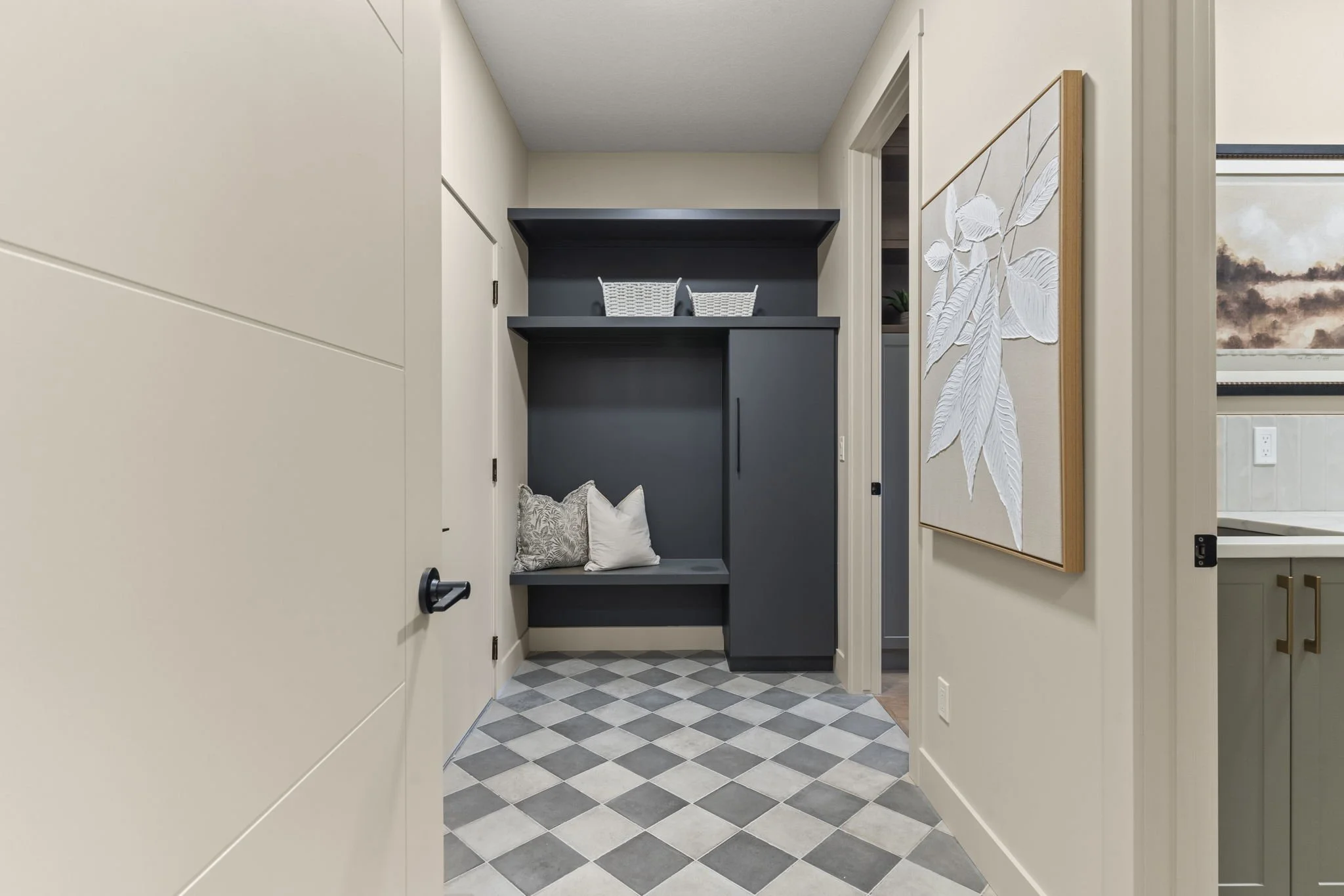 Hallway with geometric checkered gray and white flooring, beige walls, a black built-in nook with decorative pillows, white basket, and a large wall art piece with white textured leaves on a wooden frame
