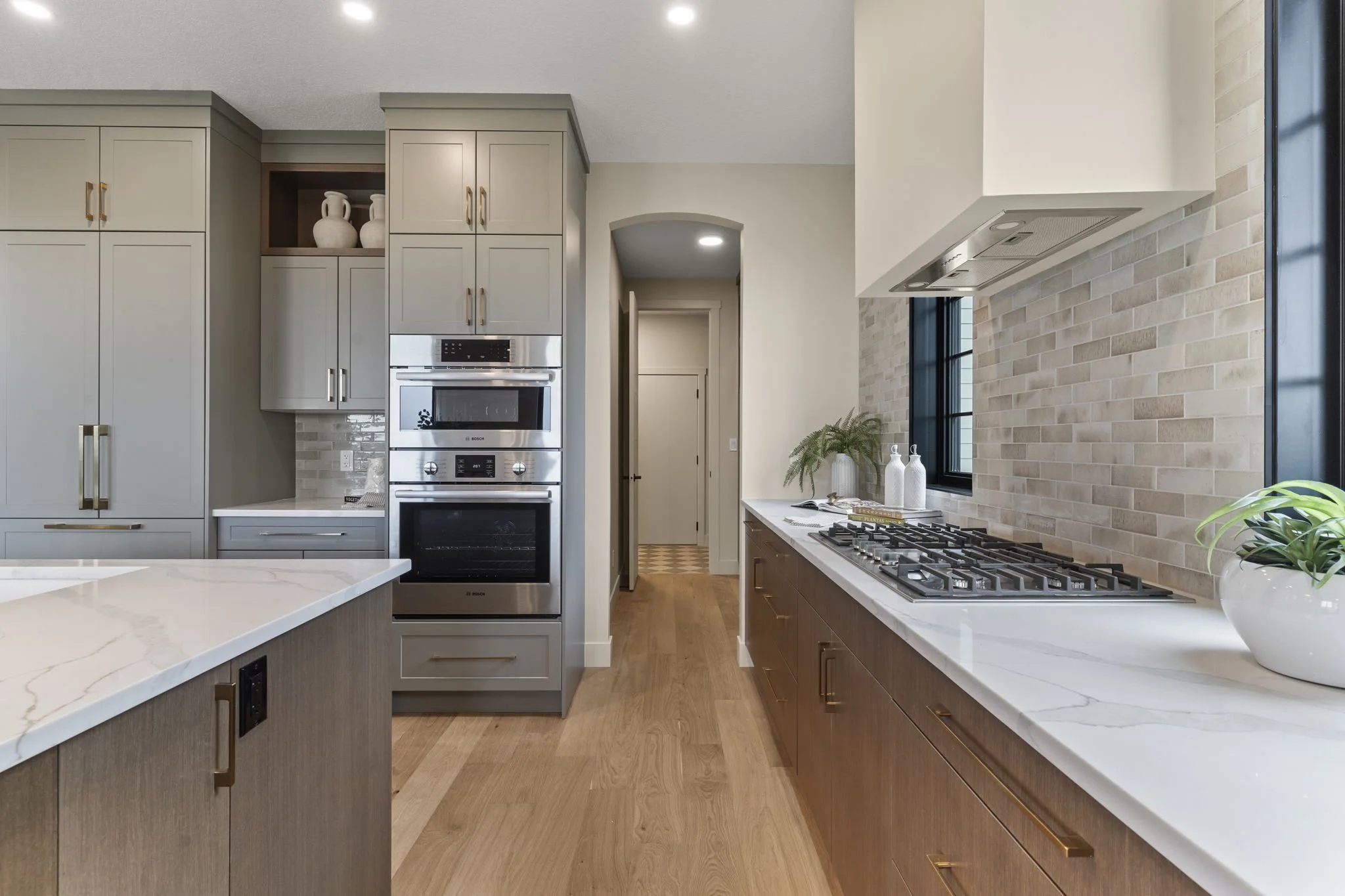 Modern kitchen with white countertops, light gray cabinets with gold handles, brick backsplash, built-in double oven, gas stove, and decorative vases with a plant by the window.