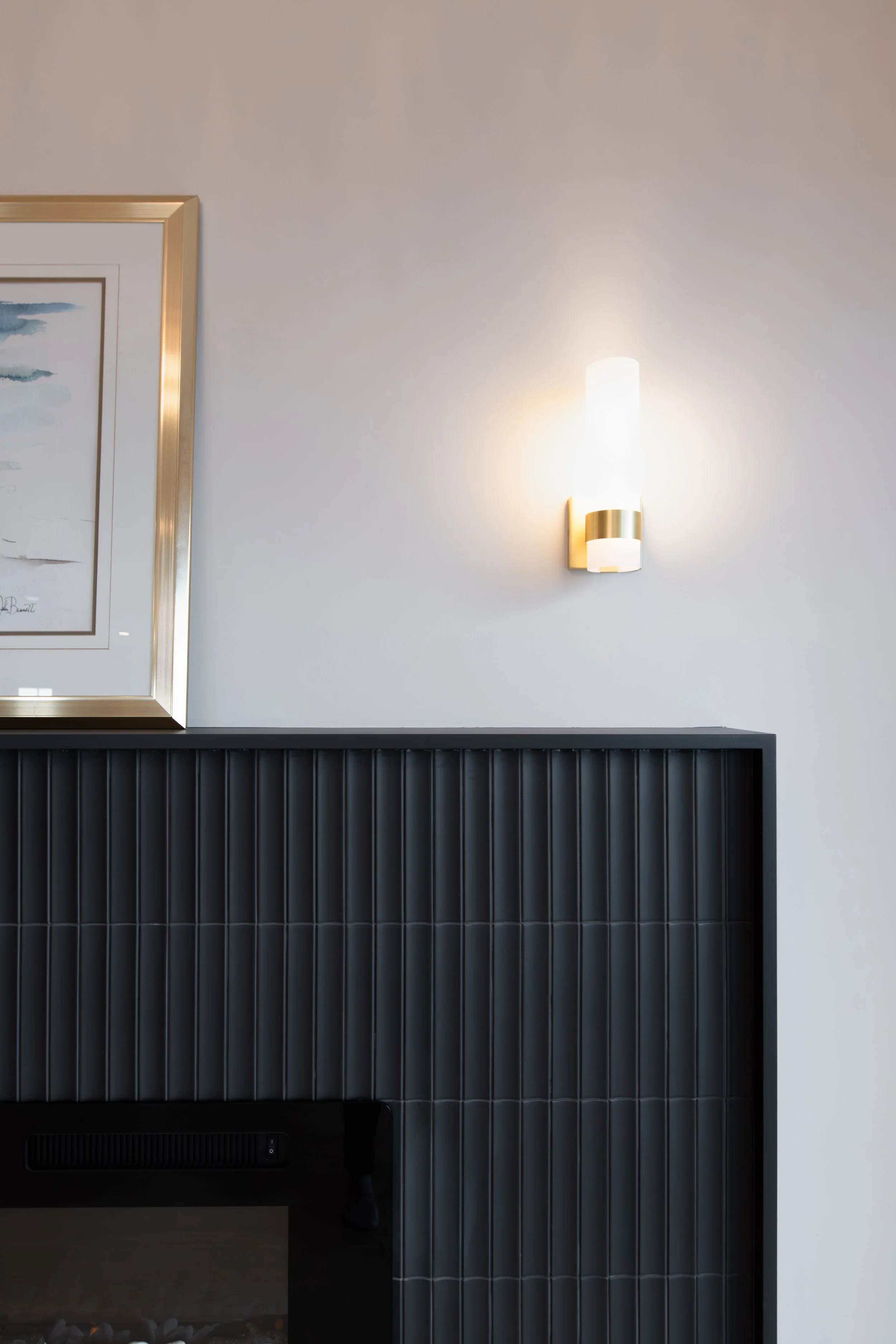 Photo of a modern wall-mounted light fixture with a white cylindrical shade and a gold accent, attached to a plain white wall. Part of a framed artwork with a gold frame is visible on the left.
