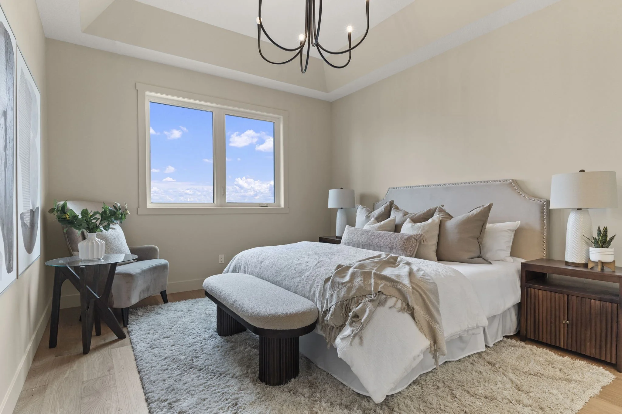 A cozy bedroom with a large bed adorned with multiple pillows and a cream-colored throw blanket, a white cushioned bench at the foot of the bed, a bedside table with a potted plant, a beige lamp, and a window showing a bright blue sky with clouds.