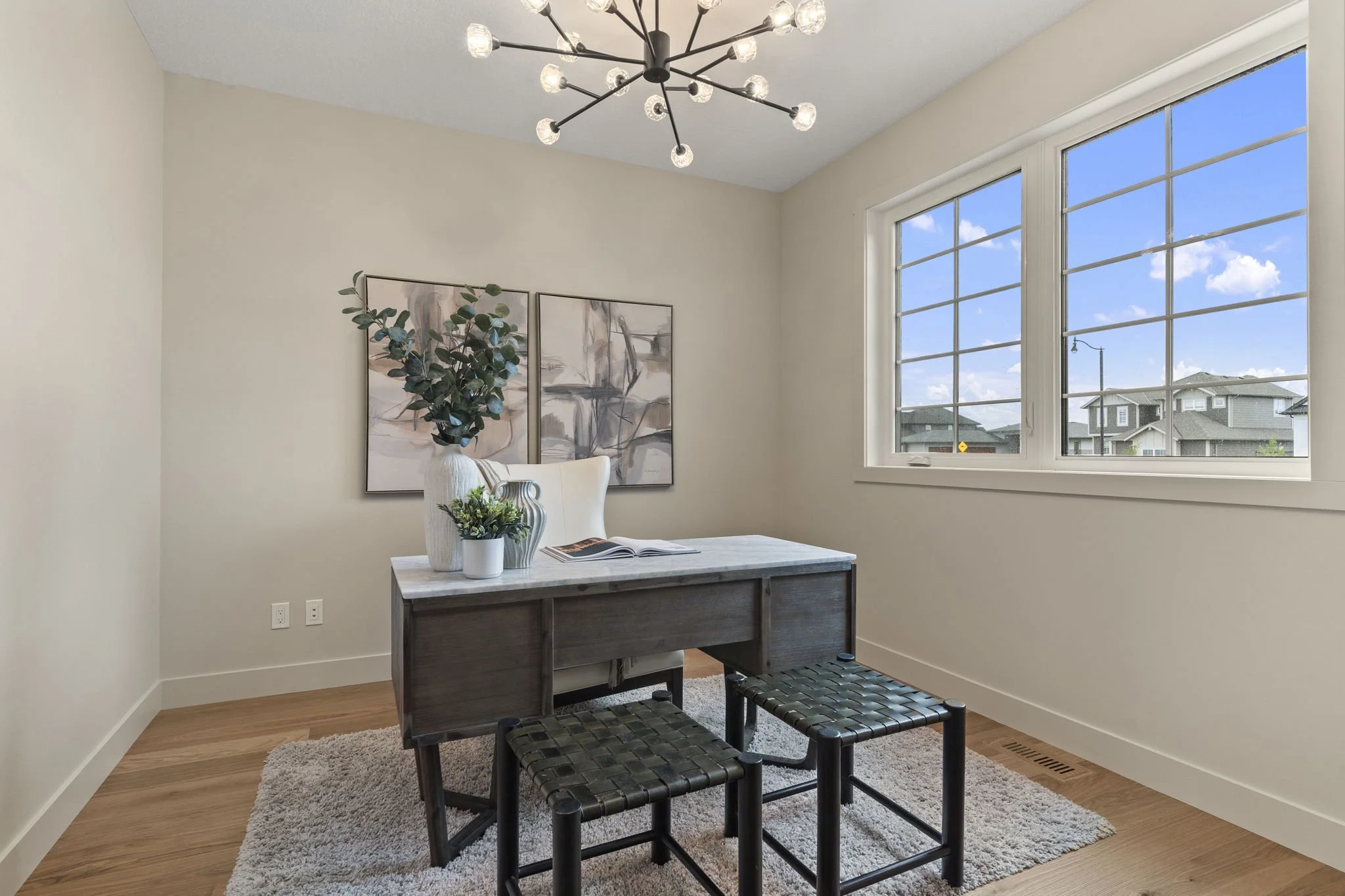 A dining area with a modern chandelier, a table with decorative vases, framed artwork on the wall, a window showing neighborhood houses, and two stools.