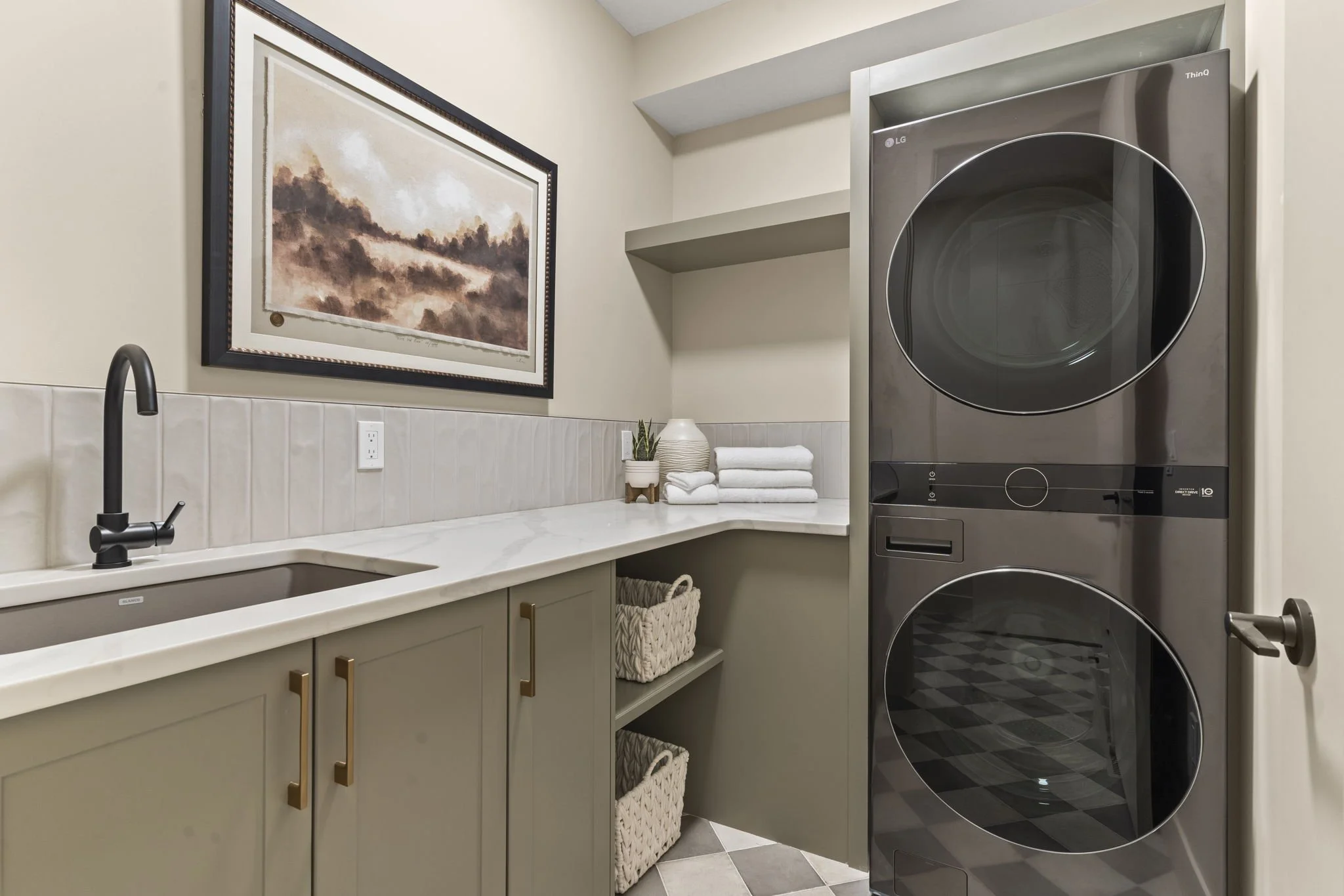Laundry room with beige cabinetry, white countertop, black faucet, framed landscape painting, stacked white towels, potted plant, and a stacked LG washer and dryer unit.
