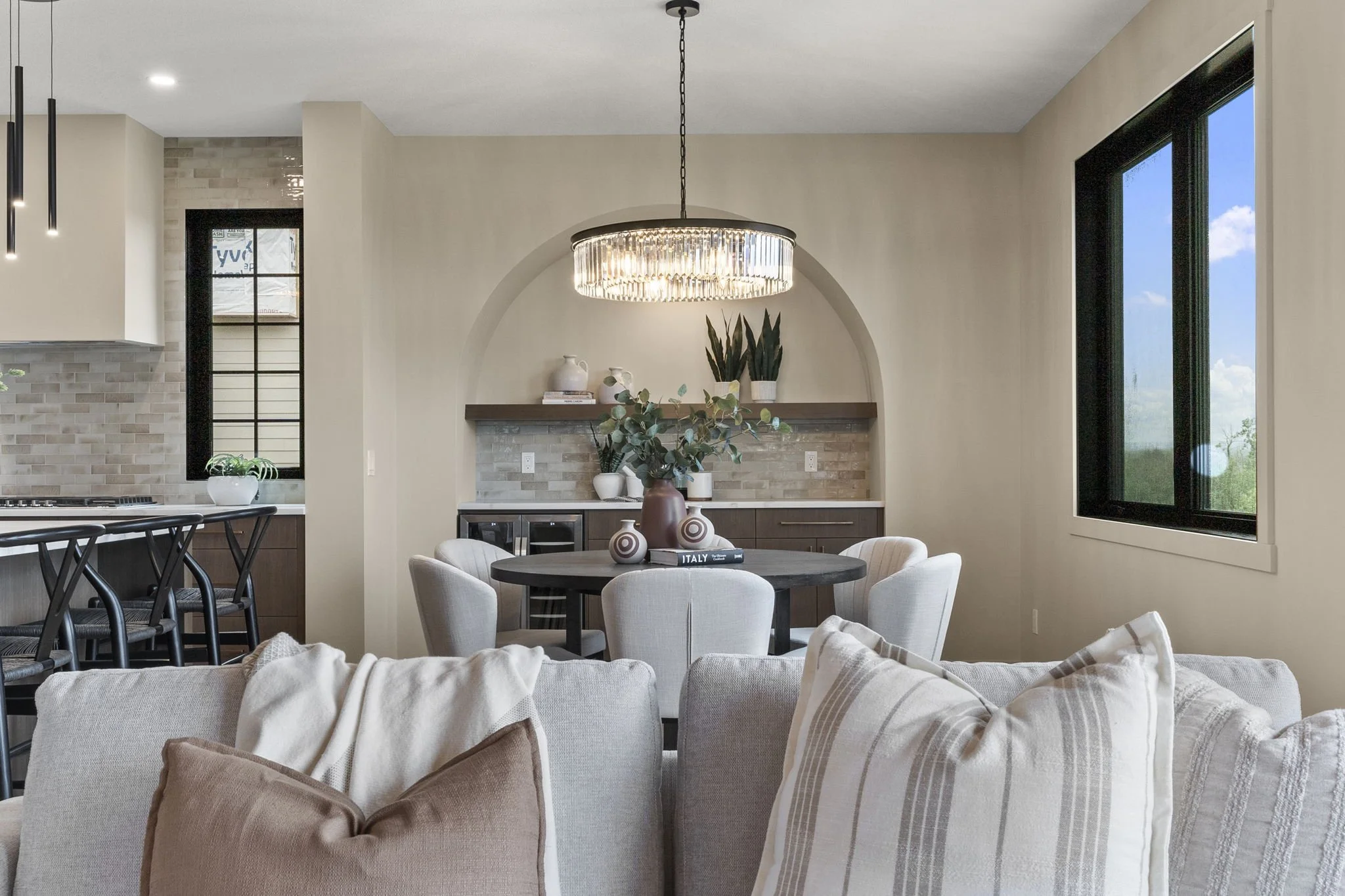 Interior of a modern living room with a round dining table surrounded by white chairs, a chandelier overhead, and large windows showing a blue sky with clouds.