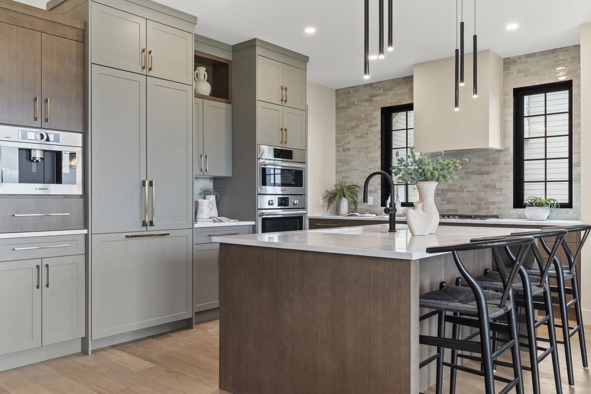 Modern kitchen with light gray cabinets, a white countertop island, dark chairs, built-in stainless steel oven and microwave, black pendant lights, a white vase with greenery, and a brick accent wall with two black-framed windows.