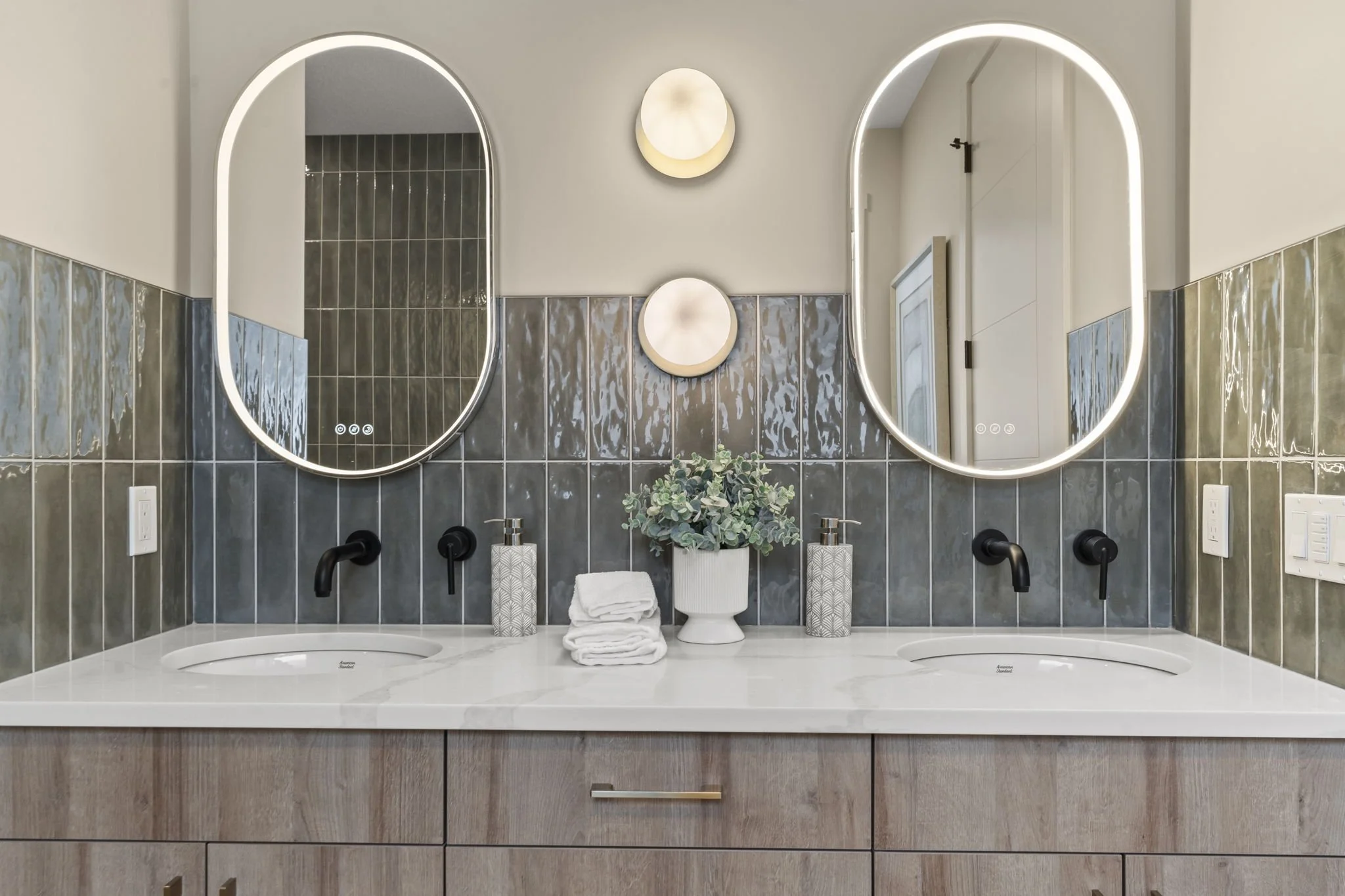 A modern bathroom vanity with two illuminated oval mirrors, a toothbrush holder, a plant, towels, and dark wall tiles.