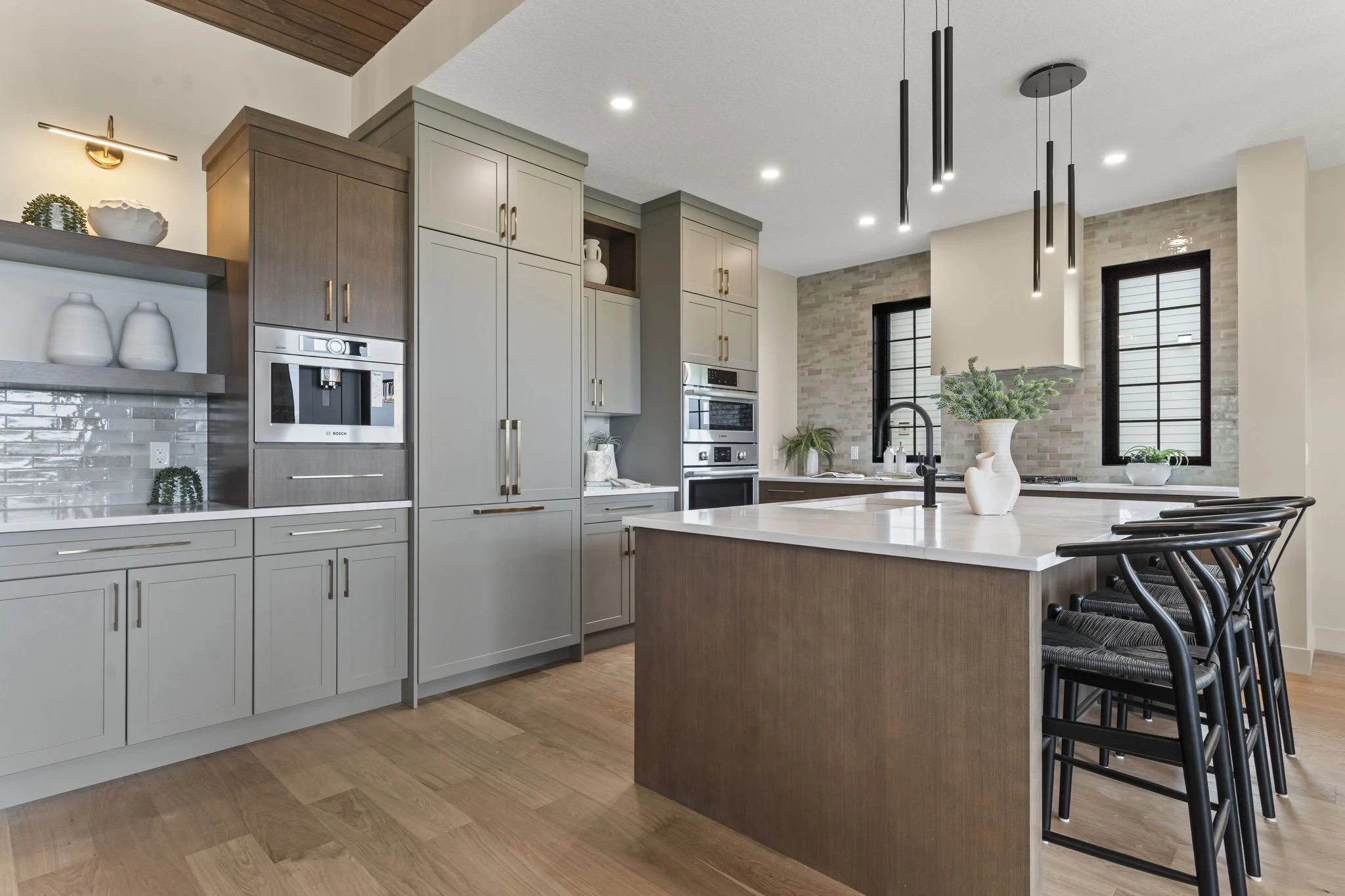 Modern kitchen with light gray cabinets, a central island with a wooden finish, black chairs, and black pendant lights. Features brick backsplash and large windows.