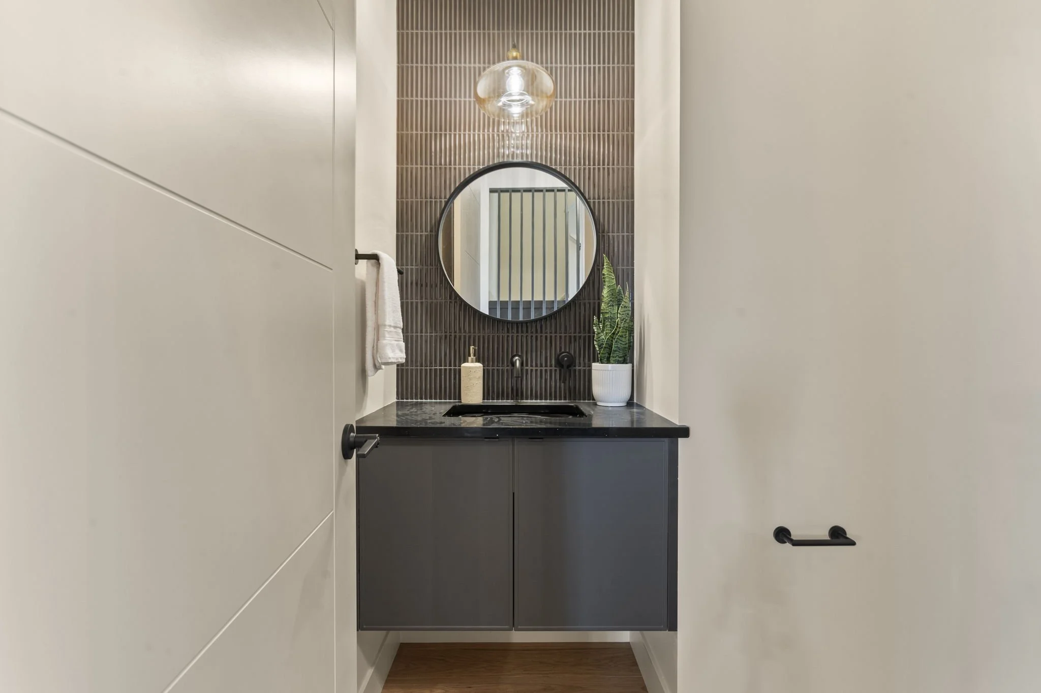 A modern bathroom vanity area with a round mirror, a black countertop with a sink, a soap dispenser, a potted plant, a wall-mounted faucet, and a textured brown tile wall backdrop.